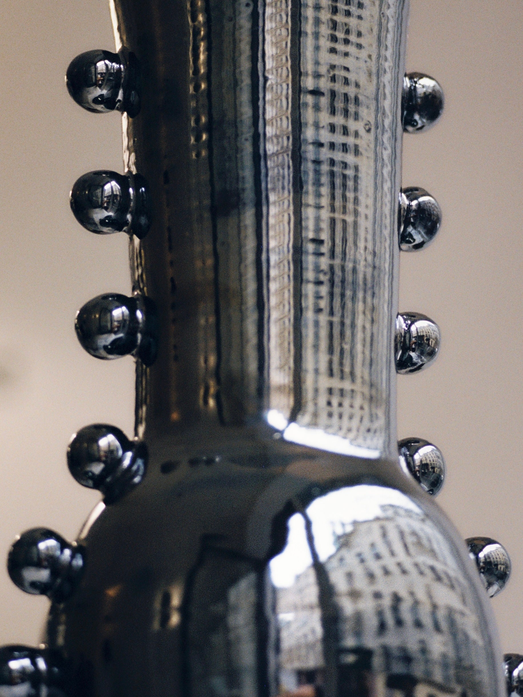 A close-up of the Vase Solstice II by Landy Rakoto Ceramics reveals its shiny metallic finish with a vertical design, adorned with signature pearls embellishing the sides. The reflective surface captures and distorts the image of a building and cityscape, showcasing its mesmerizing metallic effect.