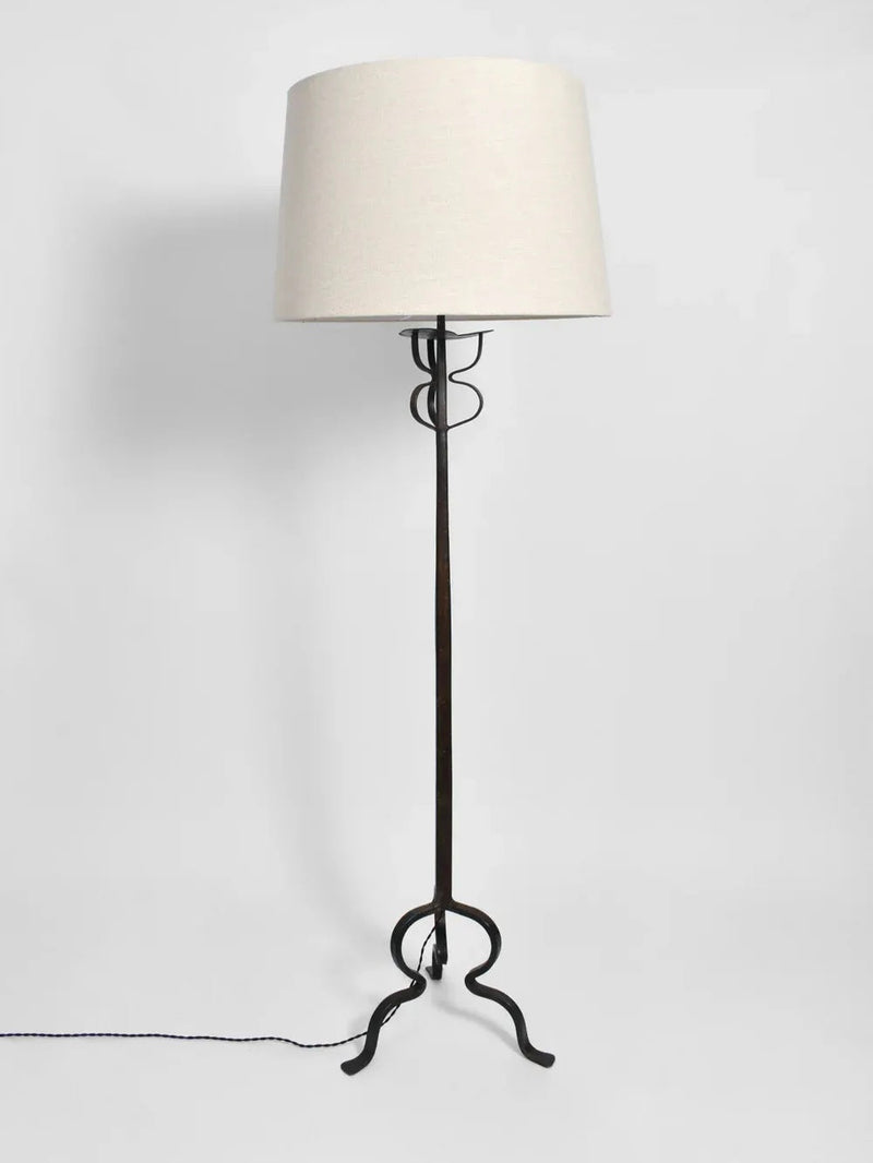 The Tripod Iron Floor Lamp, a creation of Barracuda Interiors reminiscent of 1960s France, boasts a tall black wrought iron frame with a beige cylindrical shade. Its decorative curved base and simple cord add an elegant touch as it stands gracefully against a plain white background.