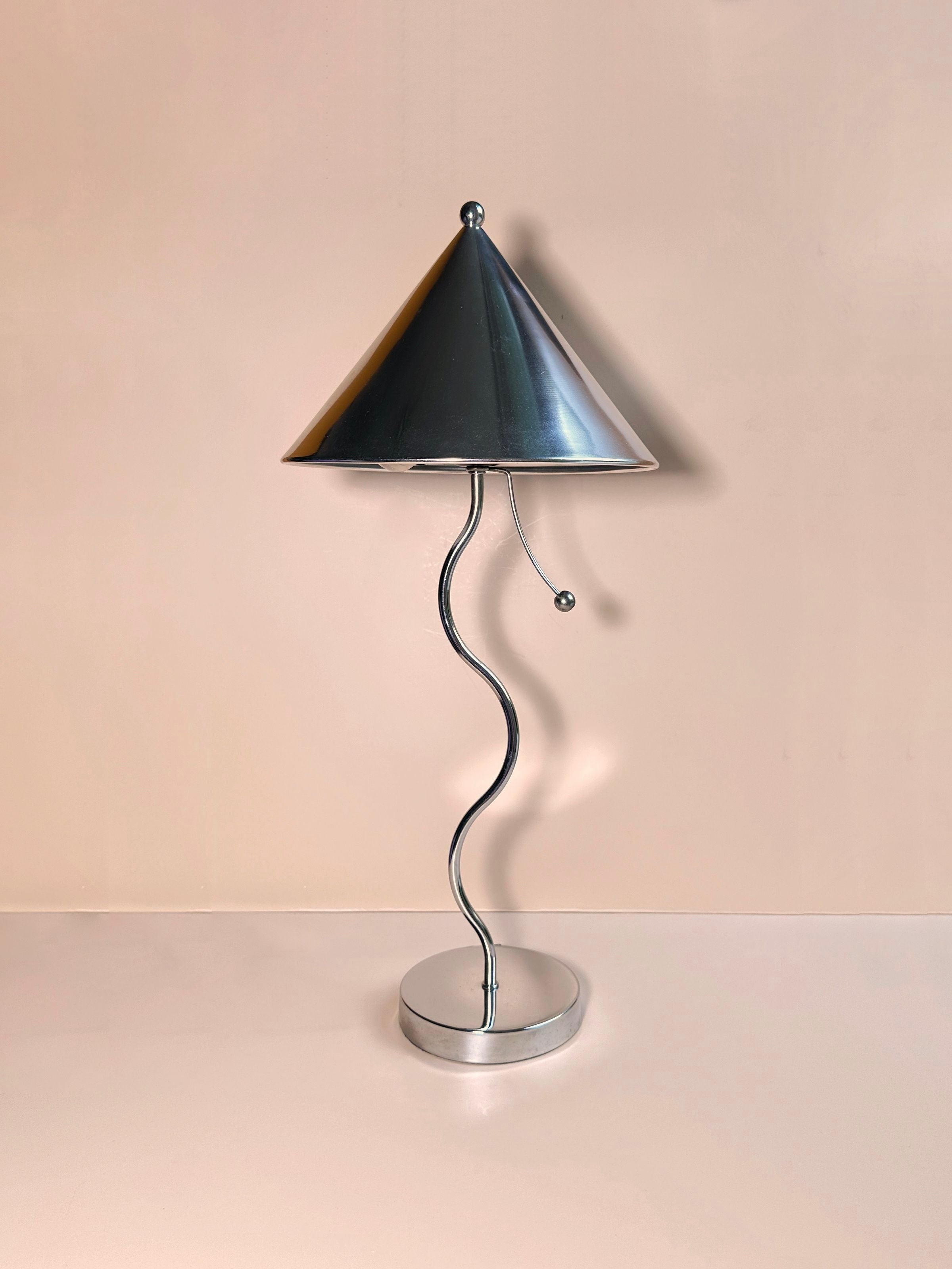 Introducing the Postmodern Wavey Chrome Table Lamp by Dodo Vintage, featuring a stylized metallic design with a conical shade and a distinctive wavy stem anchored to a round base. Crafted from chrome-plated metal, the lamp includes an innovative switch - a small metallic ball hanging from the bottom edge of the shade. Set against an off-white background that accentuates its postmodern aesthetic.