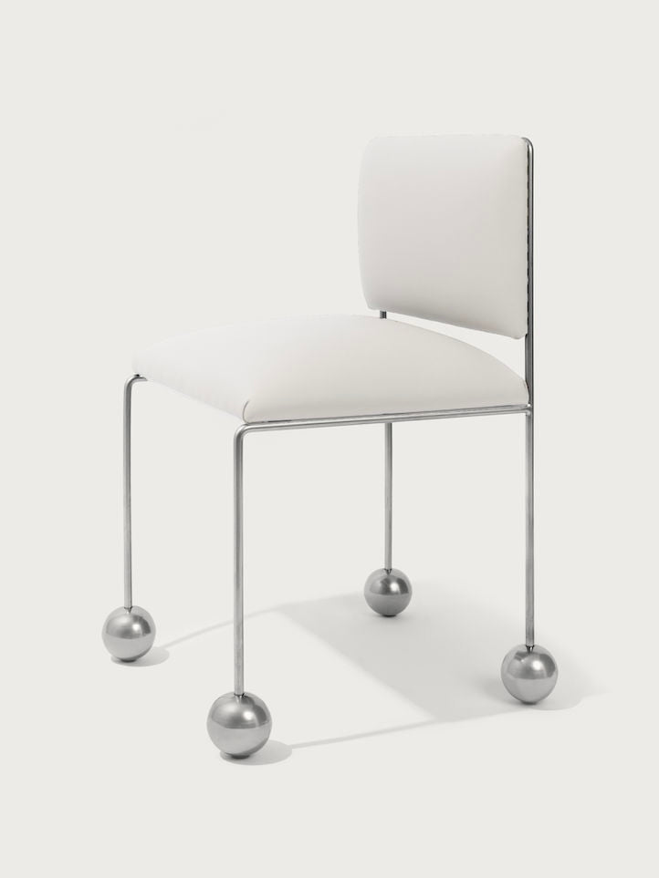 The 'Ball Foot' Chair by Panorammma features a modern, minimalist design with a white padded seat and backrest made from vegan textile. Its metal frame has straight legs ending in large, spherical feet, offering a unique contemporary appeal.