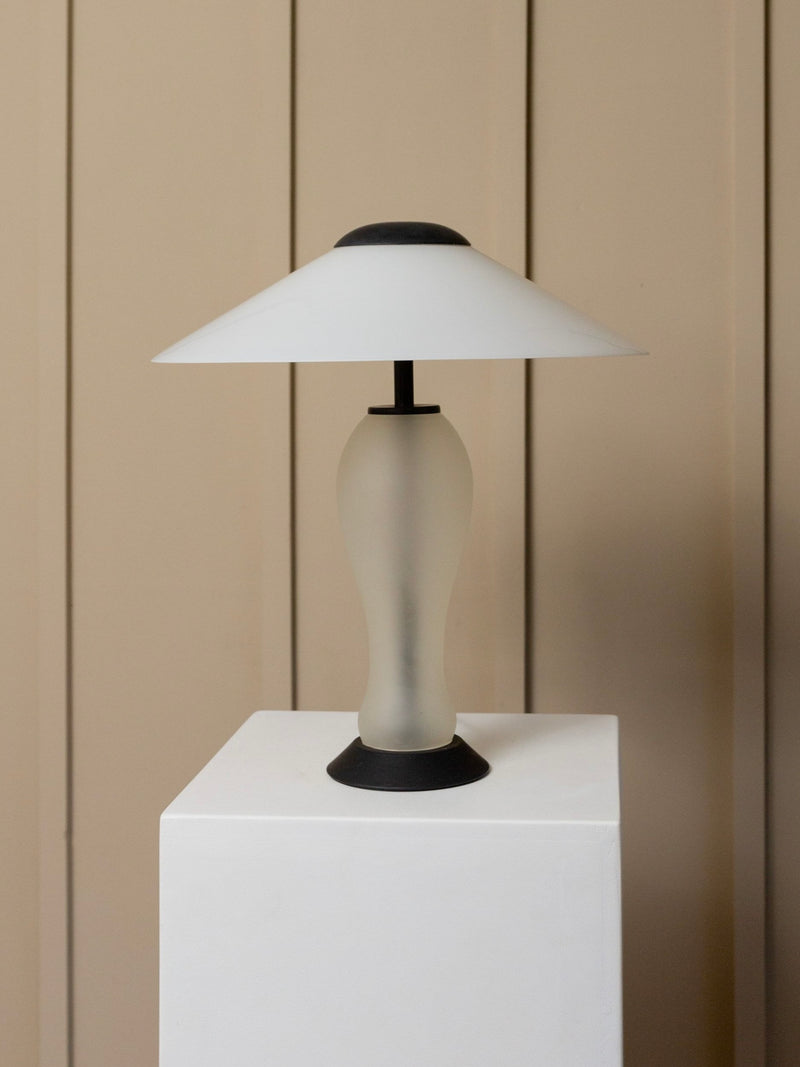 A Vintage Murano Glass Lamp from the 80s Memphis Style collection by Bottega Jacobs, featuring a frosted glass design with a wide, cone-shaped shade and black accents, sits on a white pedestal against a beige paneled background.