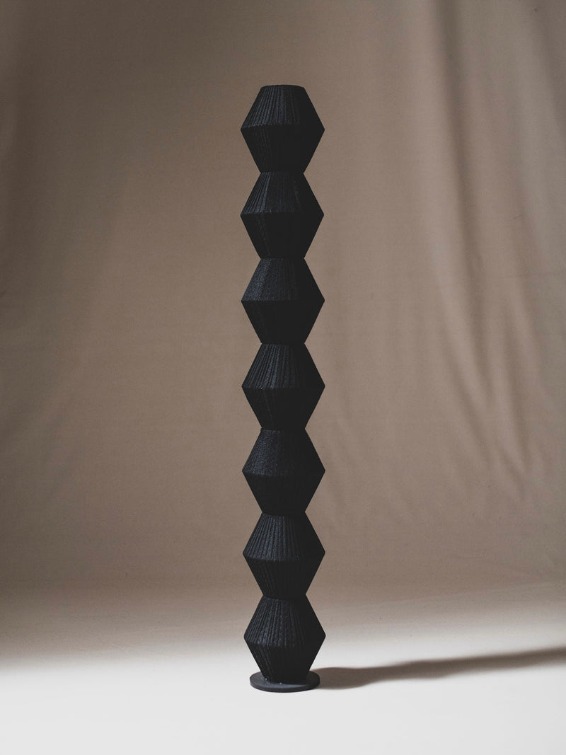 The "Column V" Sculpture by ELIURPI features stacked hexagonal black forms rising vertically on a light surface against a neutral draped background, showcasing artistry inspired by handmade Barcelona craftsmanship.