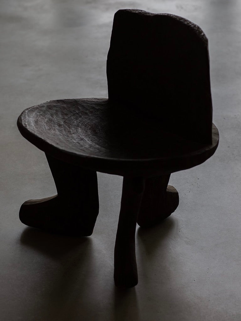 The African Stool by Bicci de' Medici features a dark, unique design with a rounded seat, thick backrest, and boot-like leg, evoking primitive cultural heritage. It casts shadows on a light-colored floor, adding an artistic touch to the minimalist setting.