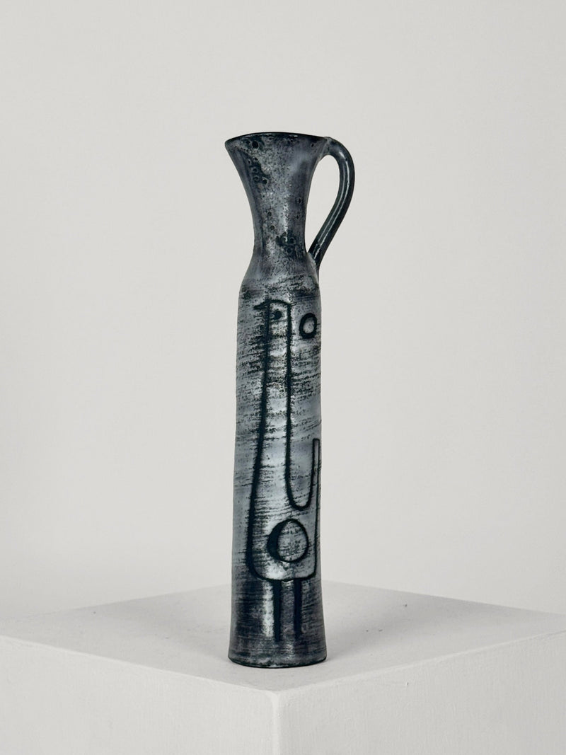A tall, slender Ceramic Bottle by Jacques Pouchain from Figuresdesign features a narrow neck and a small handle on its upper part, adorned with minimalist abstract patterns etched into its dark ceramic surface. The bottle is displayed on a plain white pedestal against a neutral background.