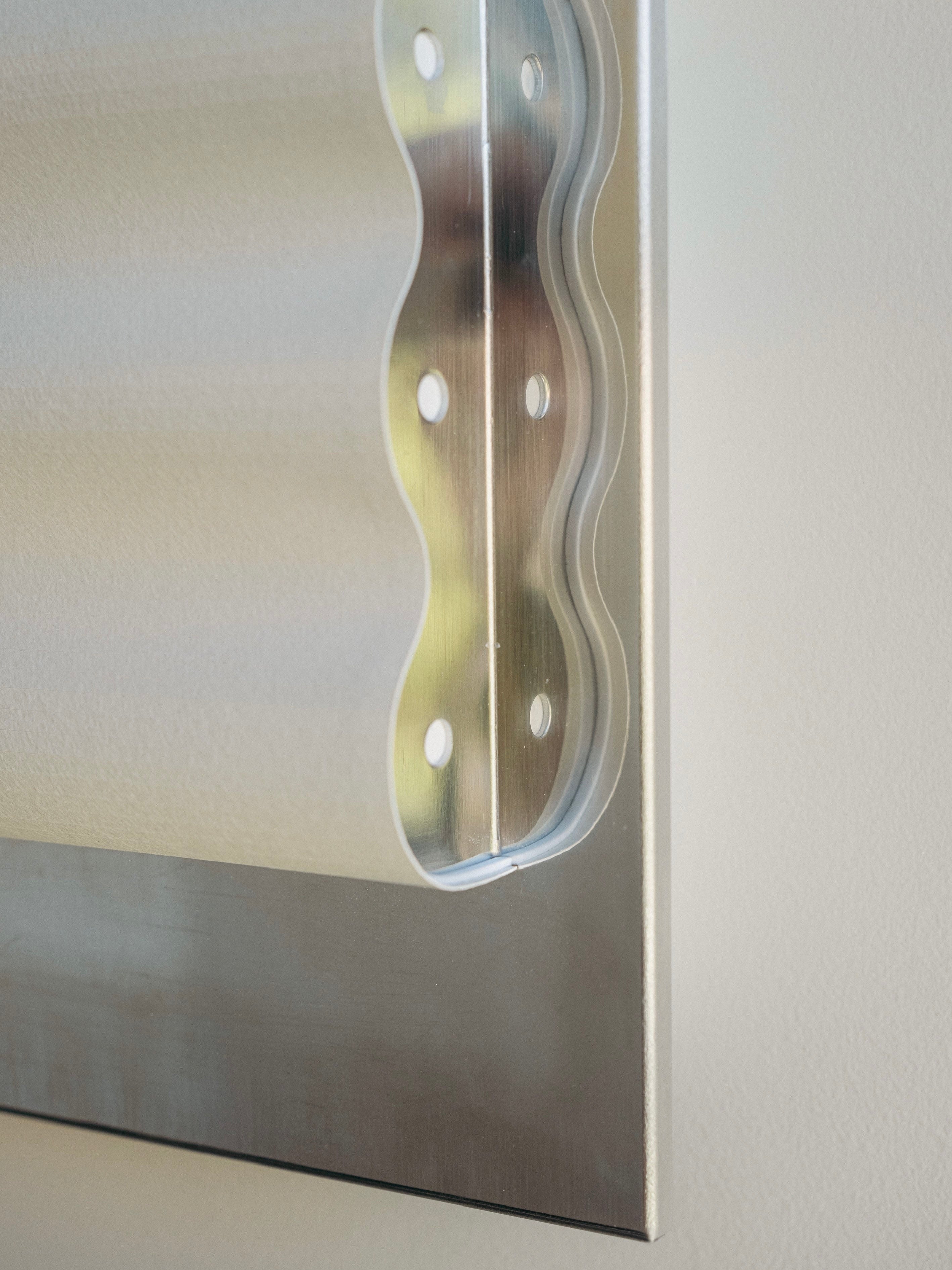 Close-up of a wavy-edged, metallic bracket from the Wall Light Frame Miroir M by Violaine d'Harcourt, featuring multiple holes, likely used for mounting or securing objects, attached to a smooth, light-colored wall. The bracket is shiny and has a modern, industrial appearance that complements minimalist design principles.