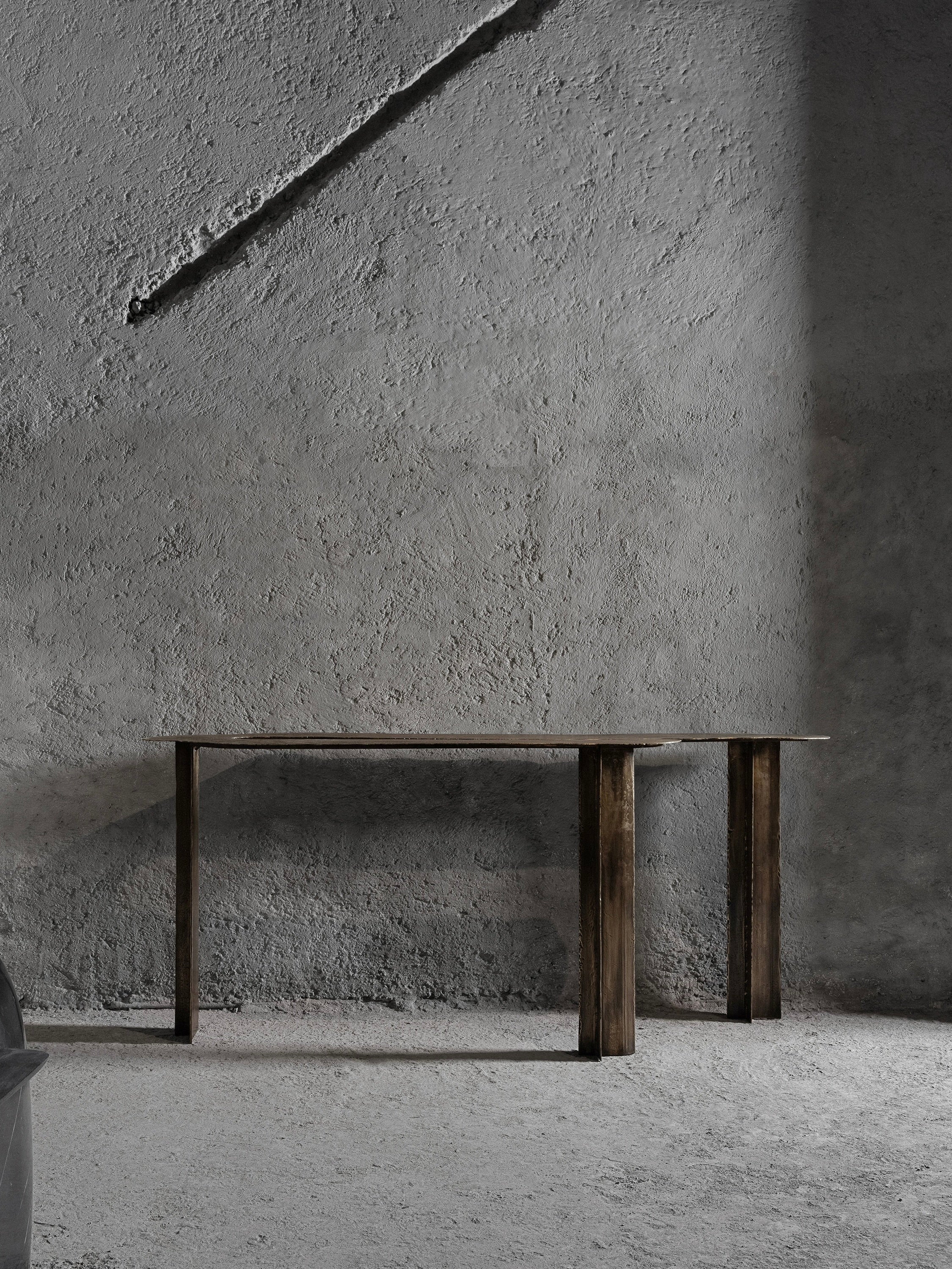 The Muisca Console Table from Ombia Studio stands against a textured gray concrete wall with diagonal shadows. The floor is concrete, and part of a round object is visible on the left, enhancing the stark minimalist atmosphere with its lighting and brass finish.