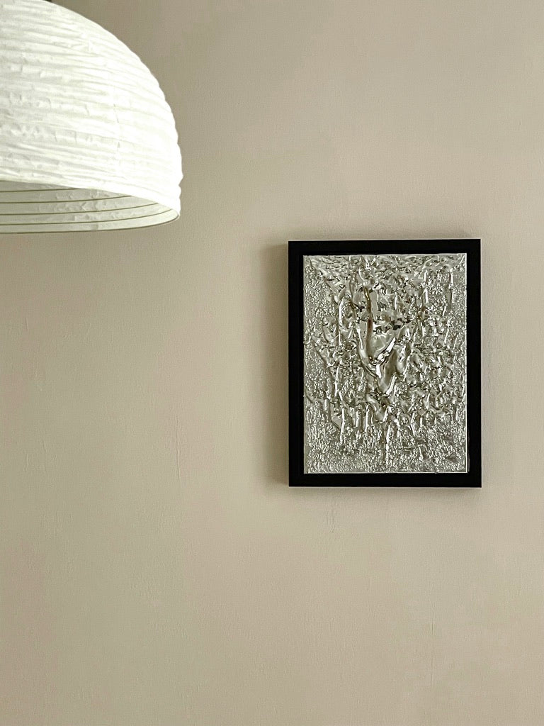 The "Almost a Mirror" sculpture by John Nordenstein, featuring a textured silver relief, is elegantly displayed in a black frame mounted on a beige wall. To the left, a large white paper lantern-style light fixture hangs from the ceiling, complementing the minimalist scene with neutral tones. Organic shapes throughout add a touch of nature-inspired beauty to the composition.