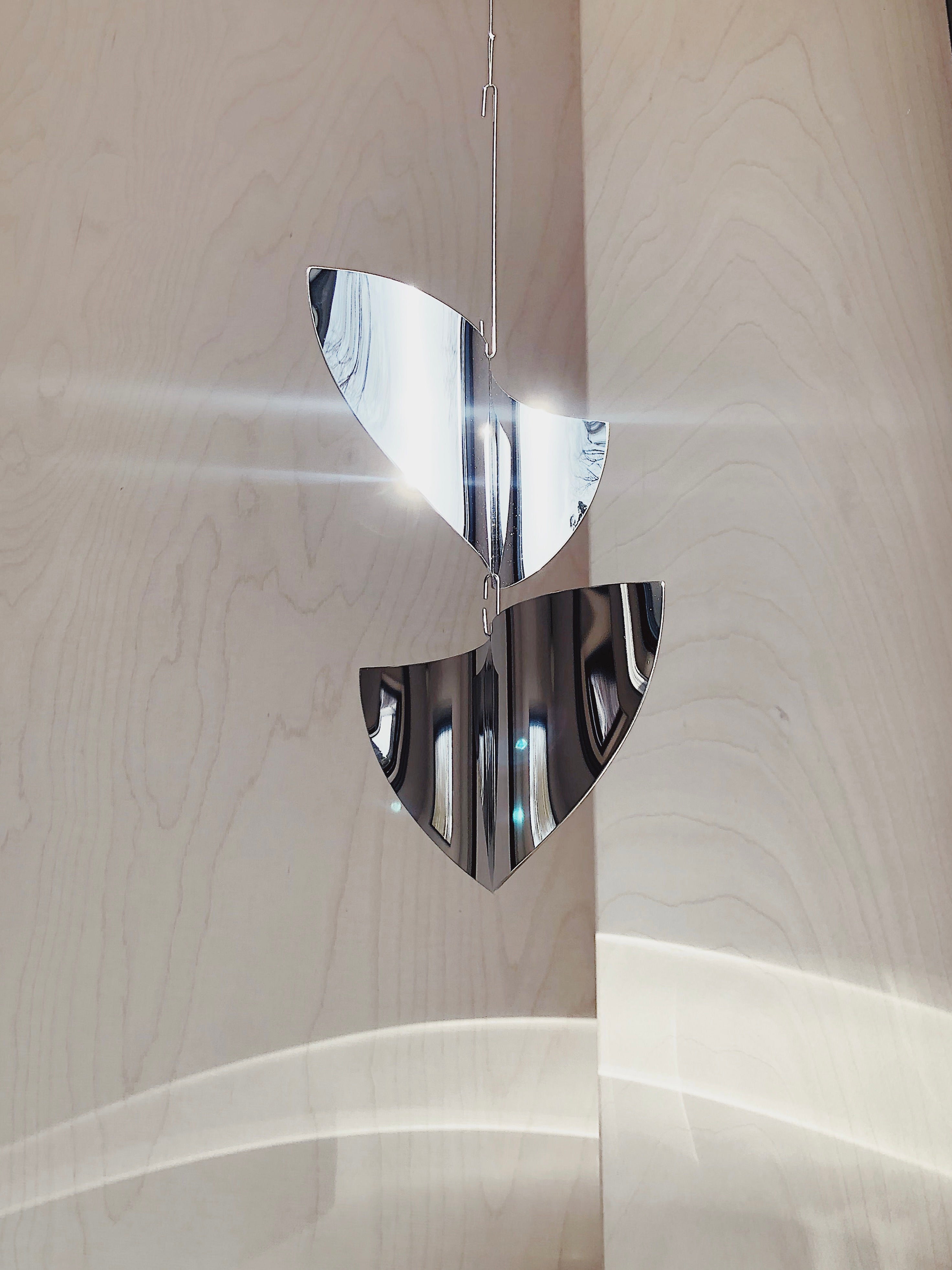 The Sun Catcher by Studio Fie Paarup features two shiny, semi-circular metallic pieces suspended on thin wire, handmade in Denmark and reflecting abstract patterns against a light wooden background.