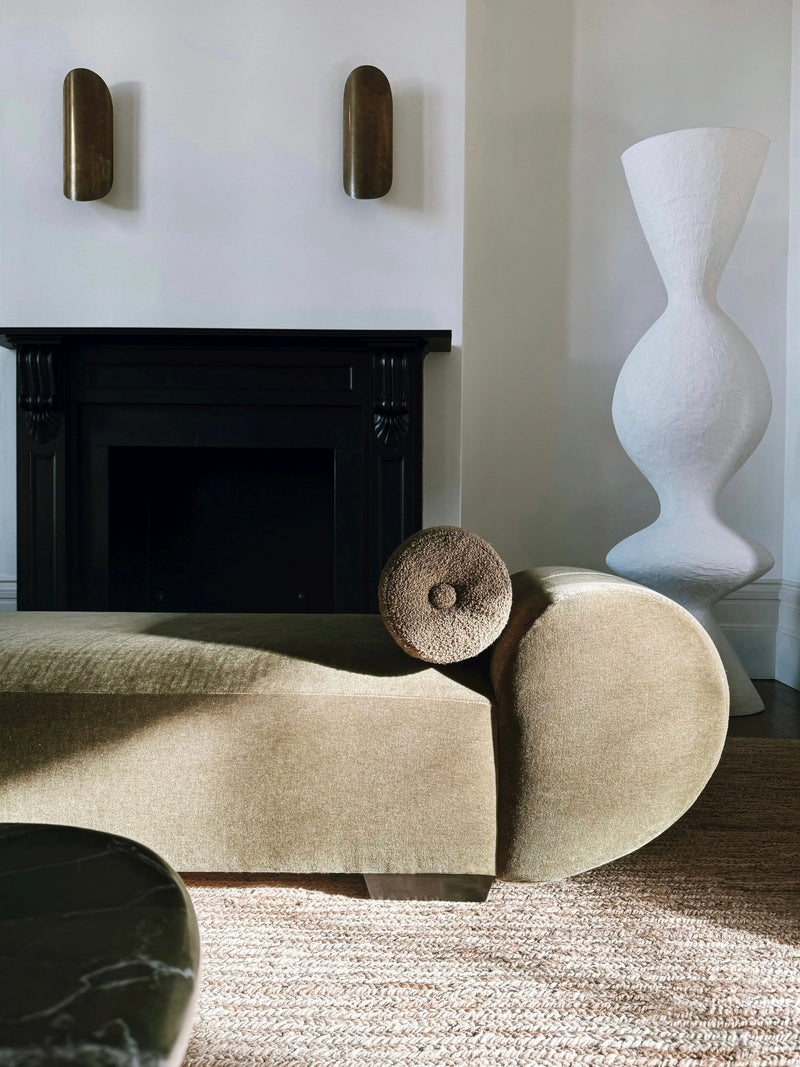 A modern living room showcases the "Sparta" Daybed by Ransom & Dunn, featuring its beige, curved design with bolster arms prominently in the foreground. Behind it, a contrasting black fireplace is flanked by two small wall sculptures. In the corner stands a tall, textured white vase, while a round dark green table is partially visible.