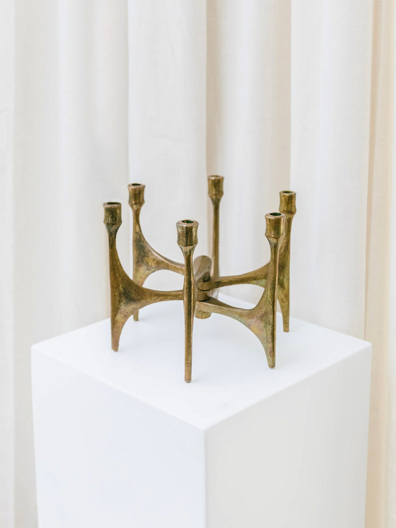 The Bottega Jacobs Candleholder by Michael Harjes in Bronze 60s, featuring a six-arm geometric design, is displayed on a white pedestal against light-colored drapes.
