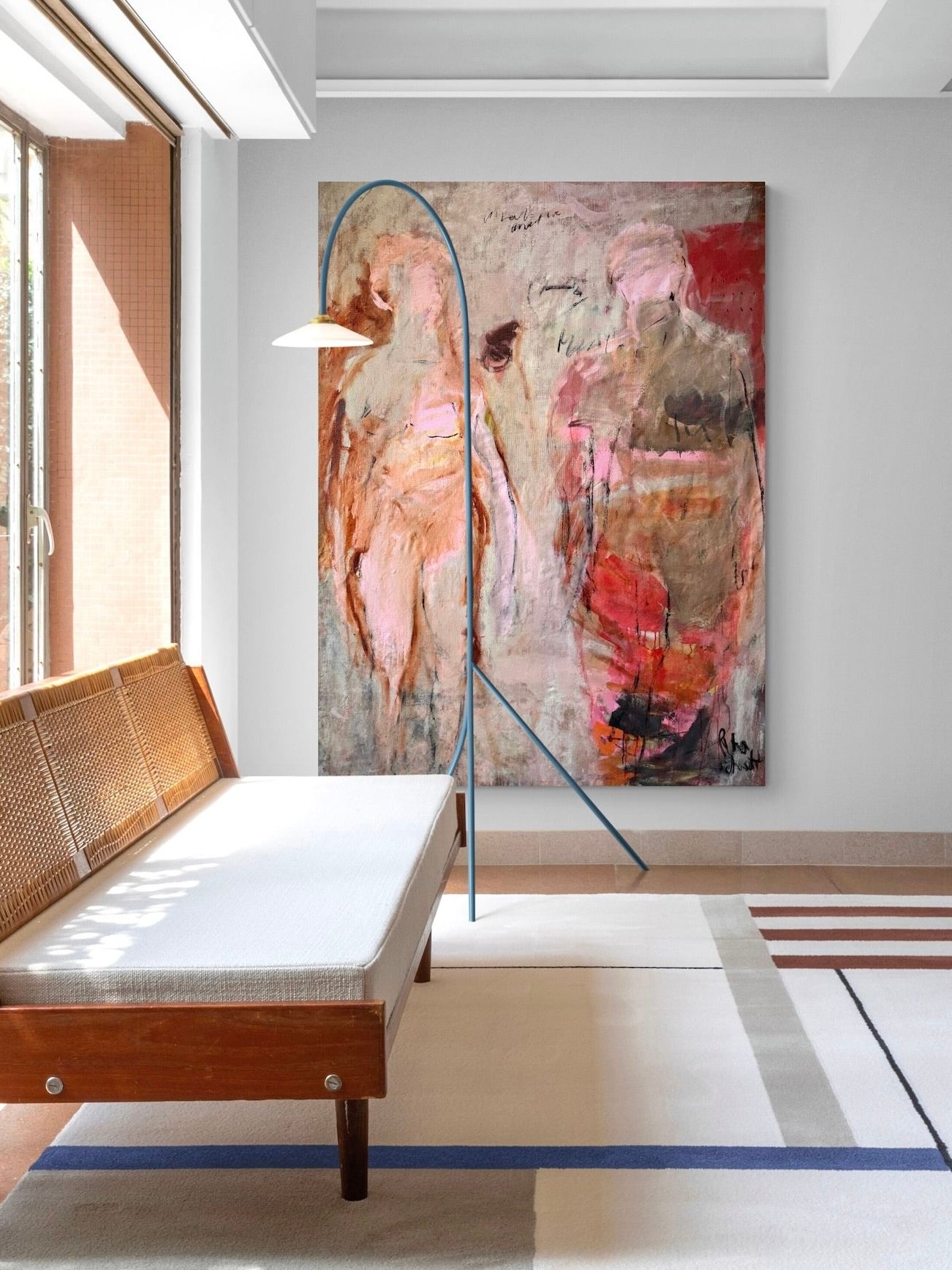 A contemporary living room exudes ethereal beauty with a wooden bench featuring a white cushion, a modern floor lamp, and Petra Schott's "What We Love" oil on canvas adding warm colors to the wall. A geometric rug covers part of the floor as sunlight streams through a large window on the left.