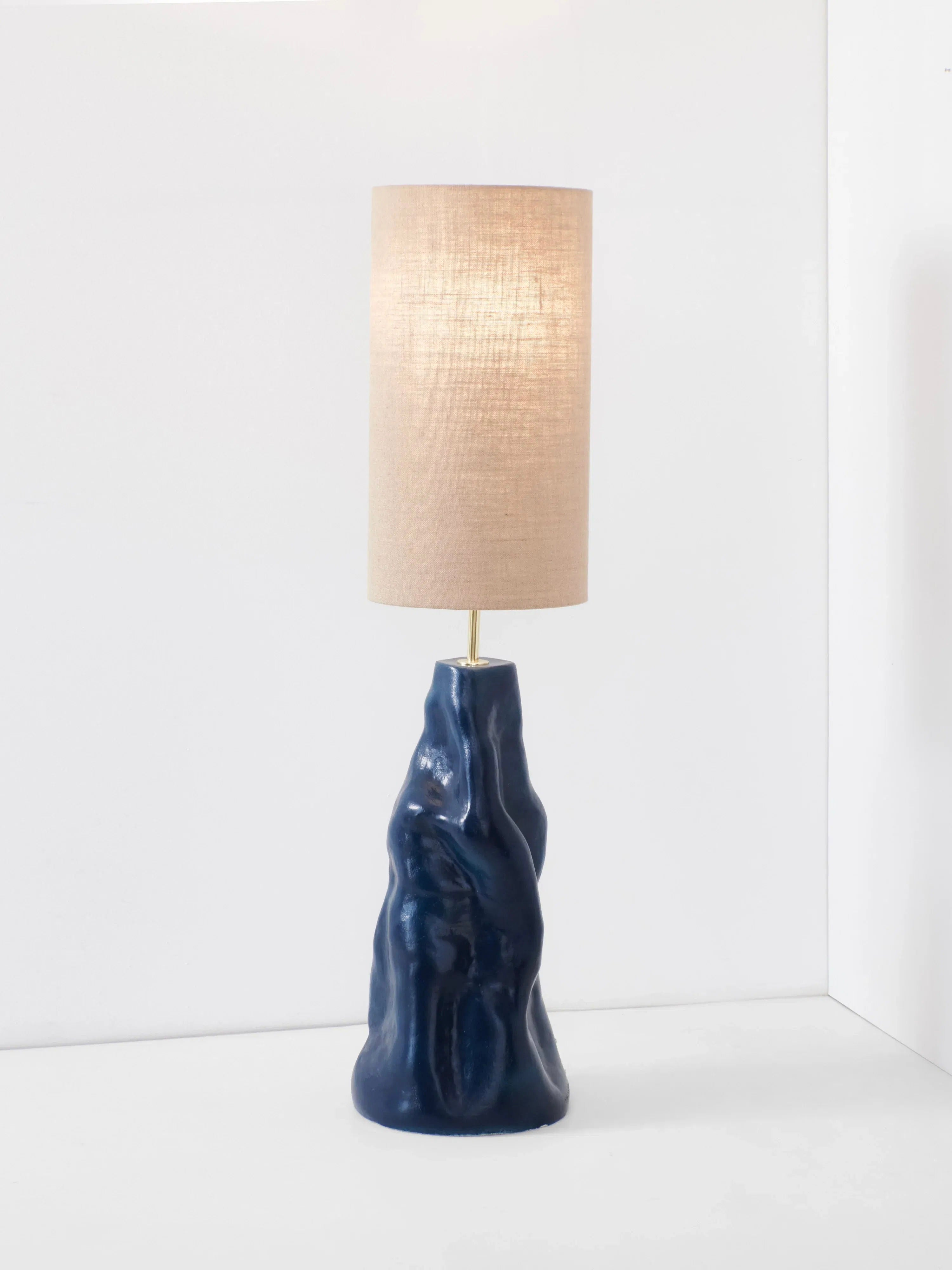 Introducing the Project 213A "Menina" Floor Lamp in Blue, a modern table lamp that boasts a sculpted navy blue ceramic base with an irregular, textured shape. Topped with a tall, cylindrical beige lampshade that offers soft, diffused light, this lamp is reminiscent of the artisanal handmade designs from Northern Portugal. It brings a chic touch to any minimalistic white room.