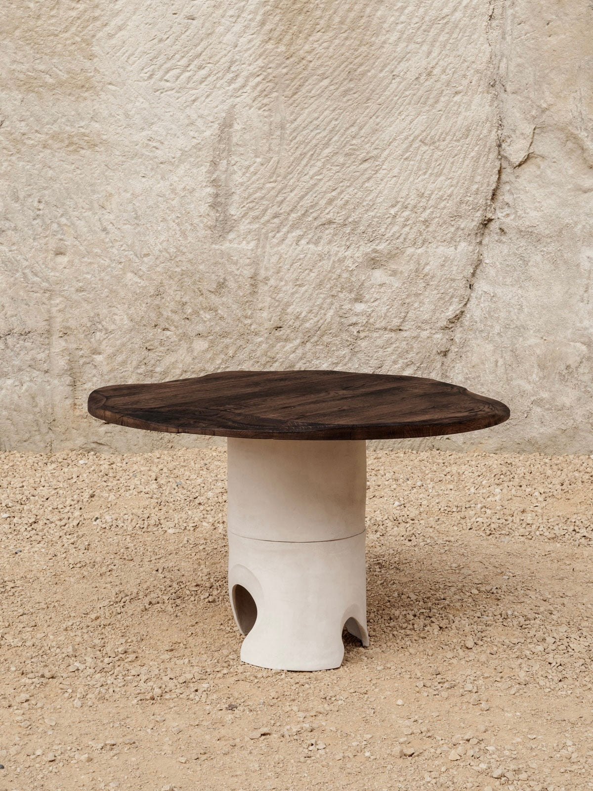 The Lac Carved Table in Sandstone and Oak by Lea Bigot features a round wooden top with a sculpted oak base, placed on a textured sandy surface against a stone-like textured wall.