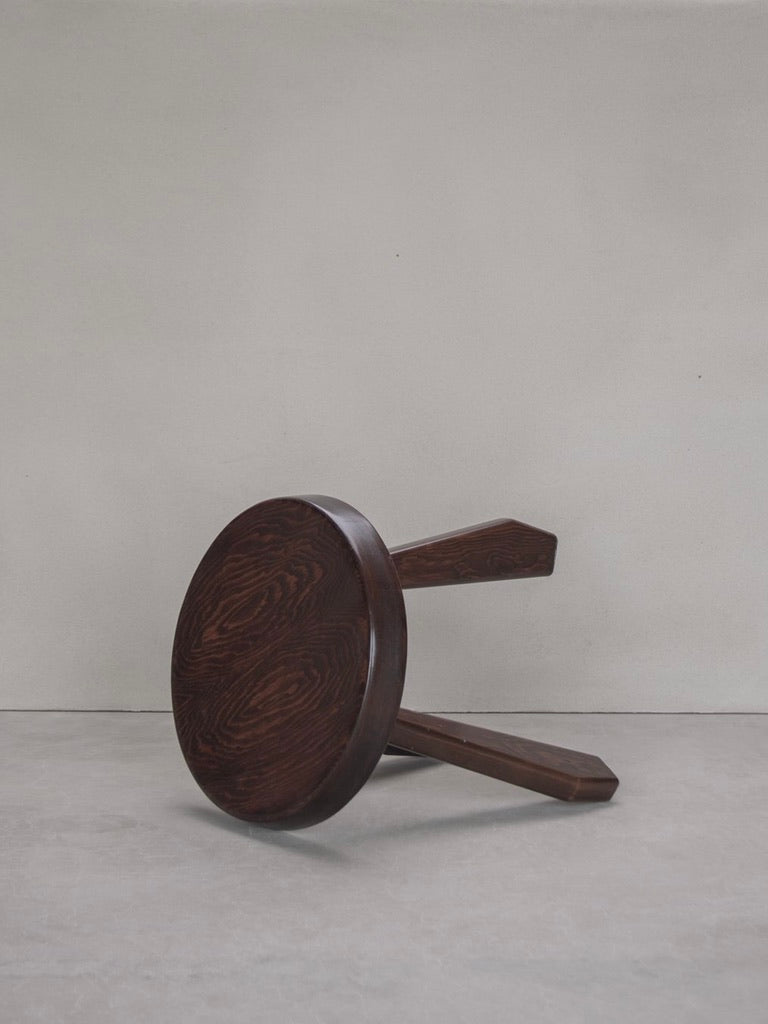 A vintage Bicci de' Medici Sandoz Stool by Charlotte Perriand, crafted from mahogany with a round seat and two visible legs, rests on its side against a plain gray backdrop, highlighting the dark finish and striking grain patterns of this timeless design.