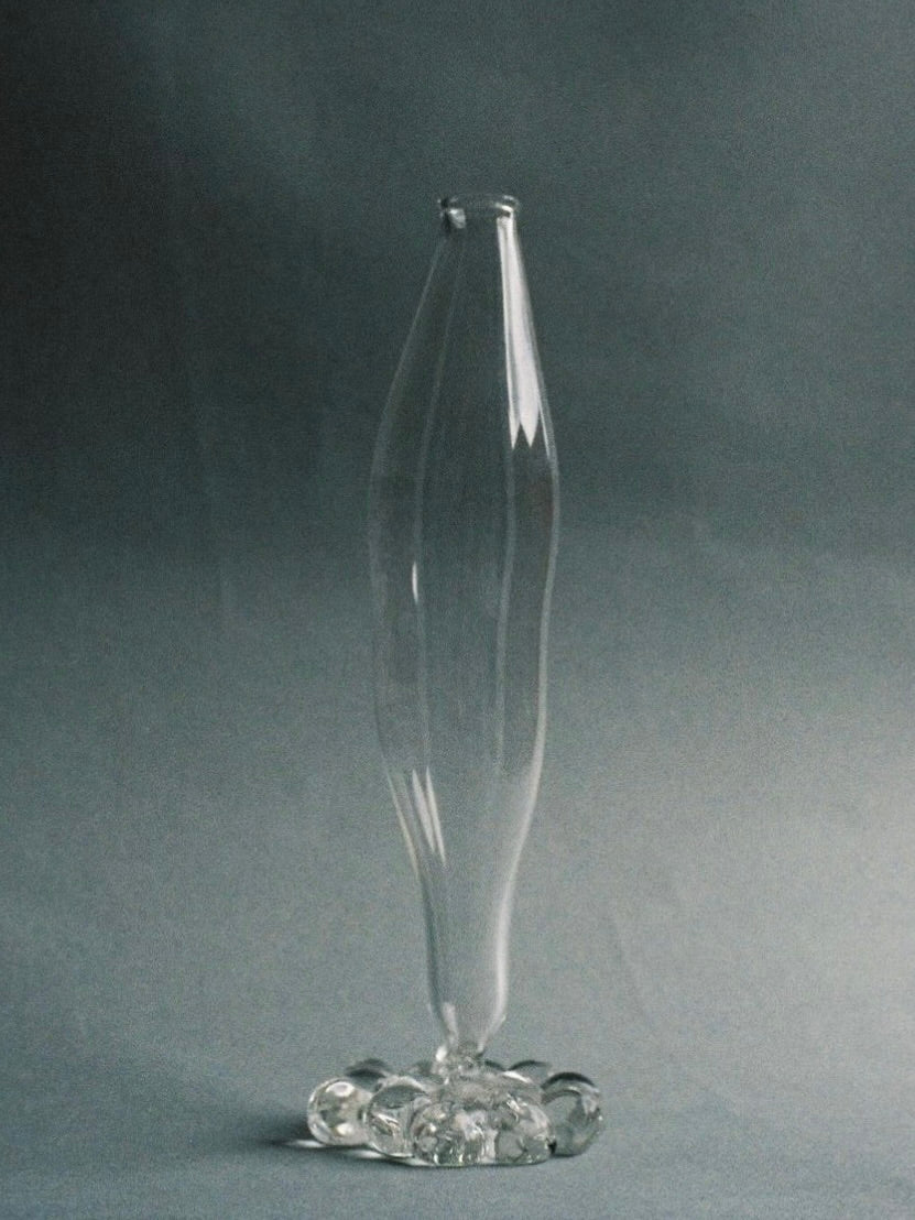 A tall, elegant Mouthblown Vase by Justine Menard stands on a flat surface, showcasing its hand-blown glass craftsmanship. The vase features a narrow, undulating body and an artistically shaped base with multiple rounded elements, giving it a unique and delicate appearance. The neutral gray background highlights the vase's transparency and long neck design beautifully.