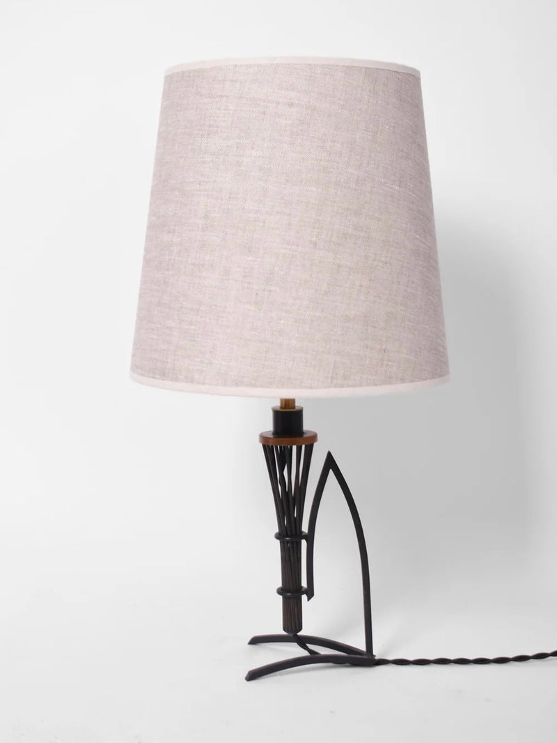 A table lamp from Barracuda Interiors, known as the Pair of Iron And Copper Lamps, 1940s, features a beige fabric shade with a decorative wrought iron base. Its design is reminiscent of 1940s France and showcases a geometric pattern against a white backdrop, while a black power cord extends from its base.