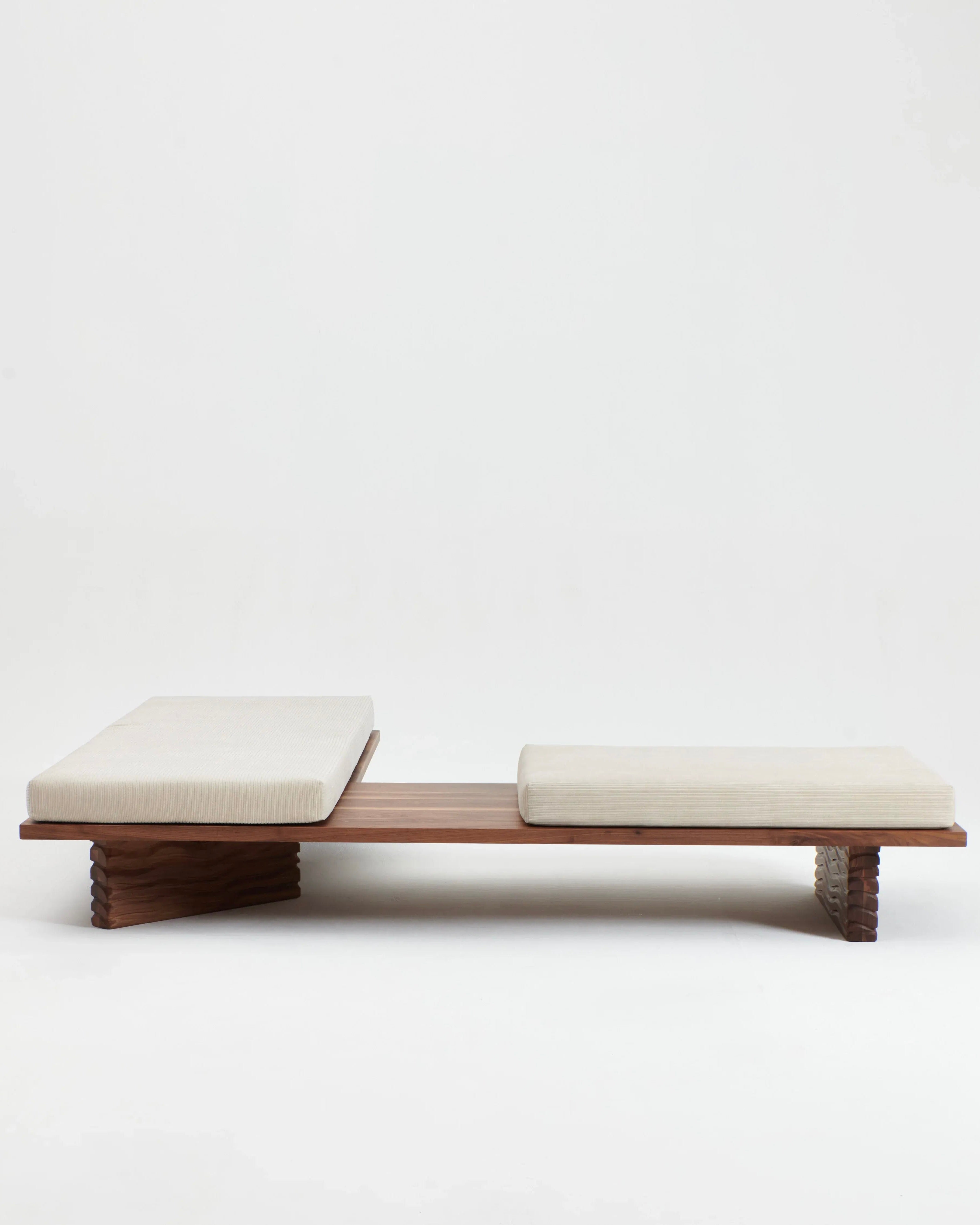 The Nazaré Daybed L Shape by Project 213A is a modern bench featuring a minimalist design with a flat solid walnut base, two neutral-colored removable cushions on top, and angled, carved wooden legs for support. Handmade in Portugal, the daybed is set against a plain white background.