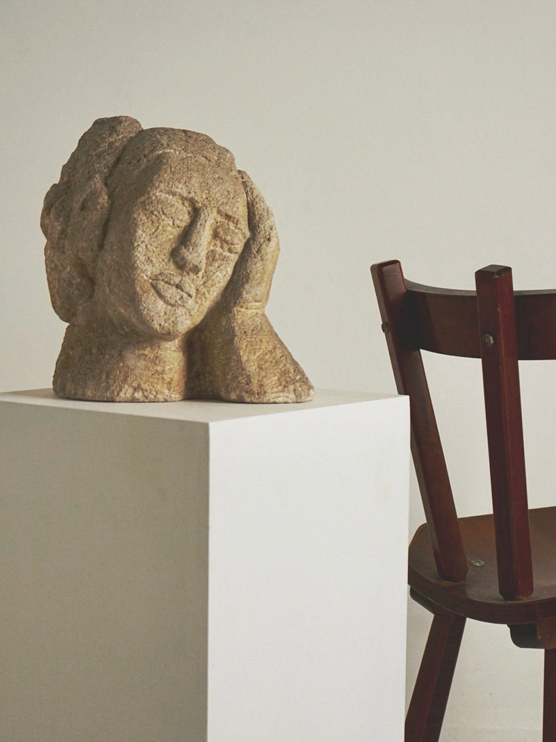The Figuresdesign Antique Carved Stone Bust of a Woman features a serene expression and rests contemplatively on a white pedestal, evoking Ossip Zadkine's elegance. Nearby, a brown wooden chair is angled against the plain wall.