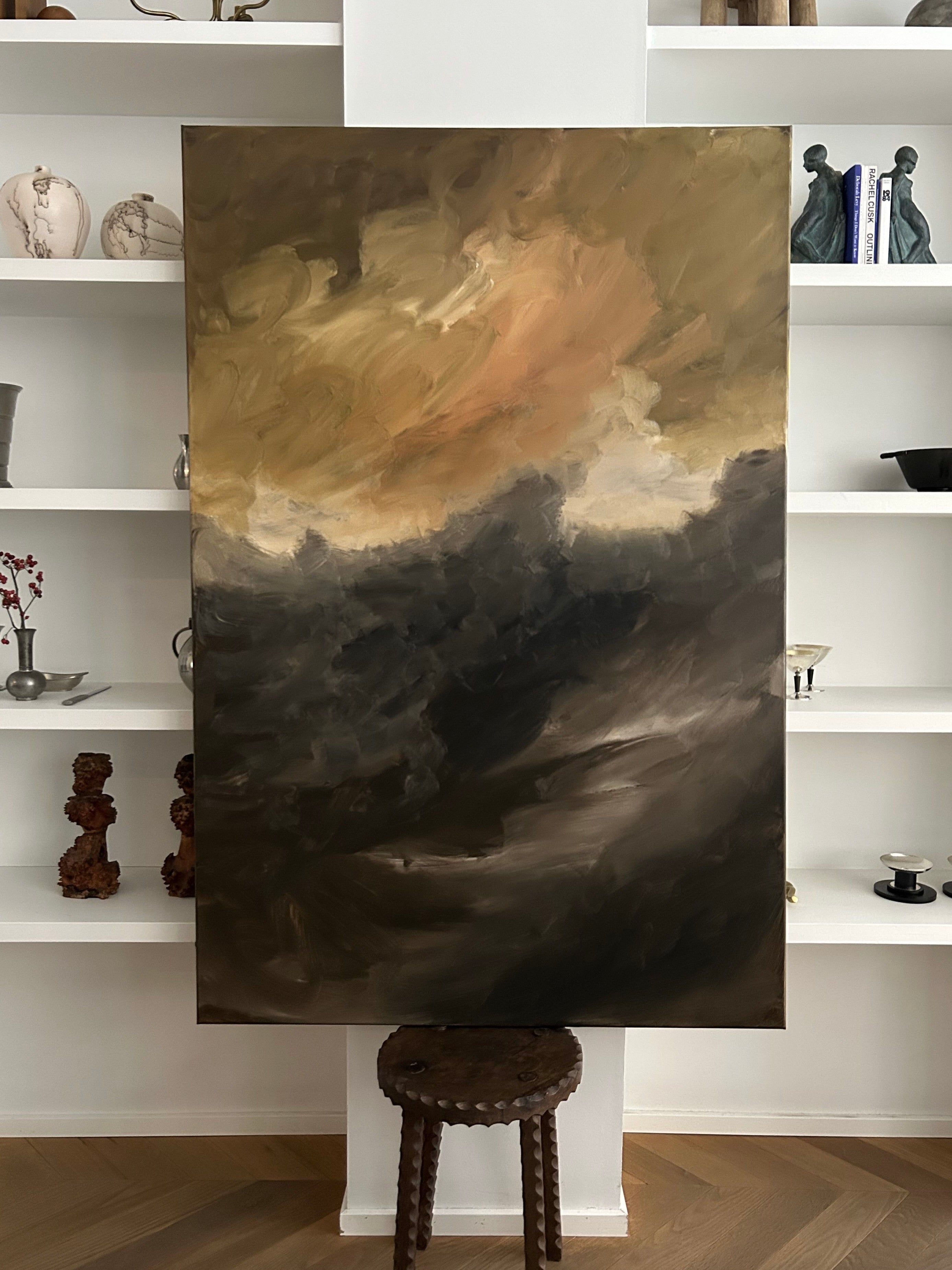 A large abstract painting titled "Whispering Peaks," with swirling dark and light tones resembling a stormy sky, is crafted by Mattias Storm. It rests on a small stool in front of white shelves filled with various ceramics and books, echoing its mixed media style.