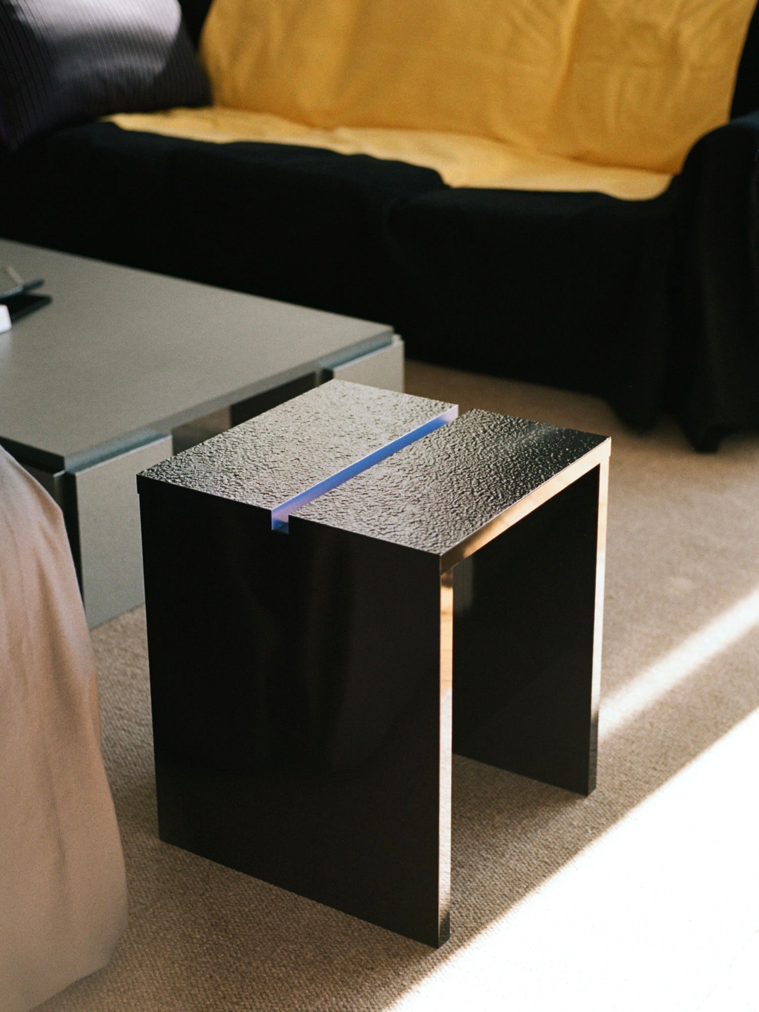 A modern living room showcases a TEGET Mara Gap Stool in wet black, set on a beige carpet. Background includes customizable furniture like a black sofa with yellow cushions and a larger gray table, contributing to a minimalist aesthetic.