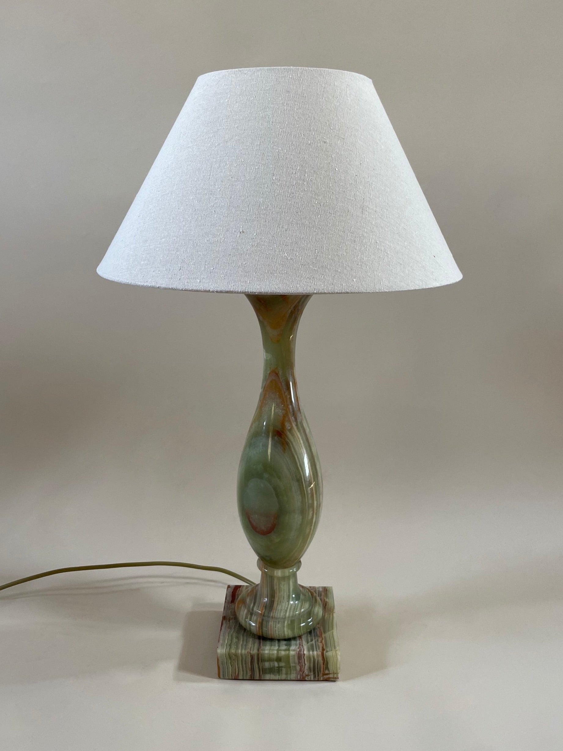 Introducing the Green Onyx Lamp C.1970 by Maud Vaughan: a vintage masterpiece featuring a white conical lampshade and a sleek marble base adorned with green and brown hues. The lamp boasts a slender, vase-like stem, complemented by an antique gold flex power cord visible at the base. Set against a neutral background with no discernible details, this piece exudes timeless elegance.