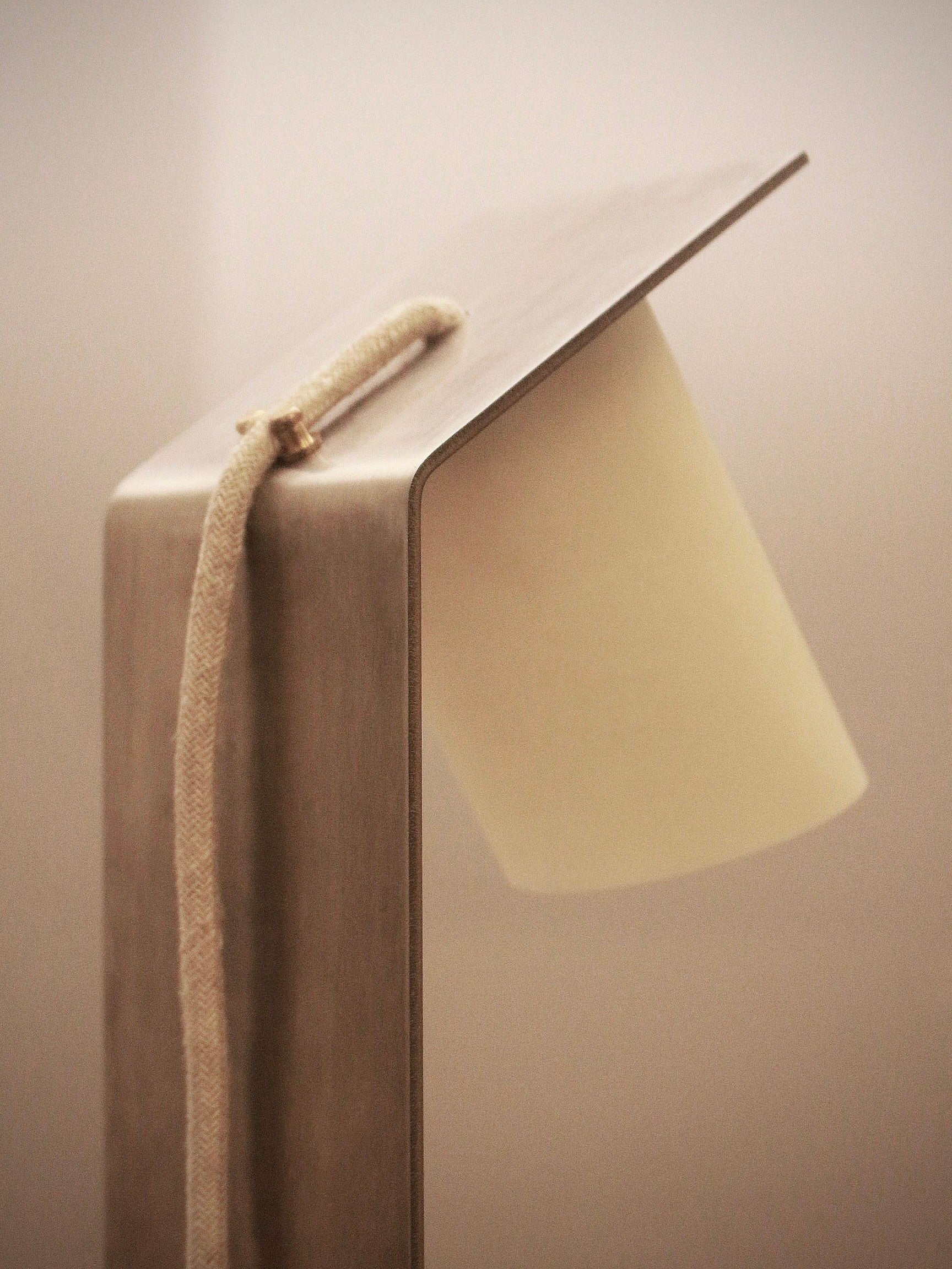 Introducing the "Bastro" Table Lamp by Pedro Avila: a minimalist, modern piece with a brushed metal finish and an angled rectangular shade. Evoking the essence of brutalism, it boasts a fabric-covered power cord that elegantly drapes over the top, perfectly blending with its contemporary design.