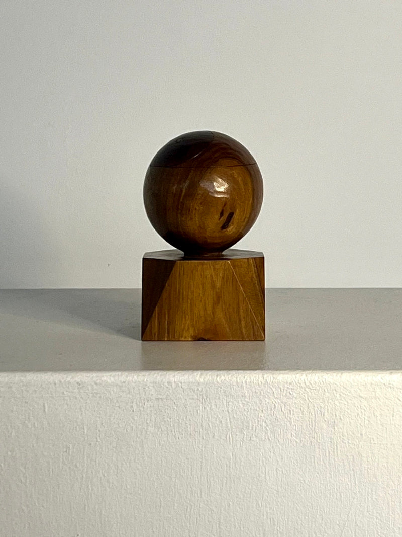 The Yin Yang Orb sculpture by Philippe Gilbert, a creation from 1982 under the Porter Studio brand, showcases a polished wooden sphere positioned on top of a square wooden base. It is displayed on a smooth, light-colored surface against an unadorned background, casting a gentle shadow.