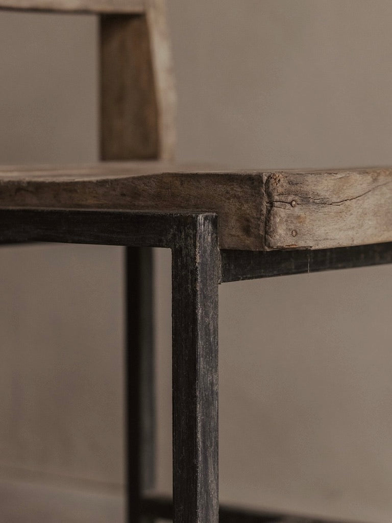 A close-up of the Bicci de' Medici's Pierre Jeanneret authentic PJ-SI-65-A chair showcases its rustic unfinished textures, with teak wood slats on the seat and backrest and metal legs, offering a minimalist industrial look against a neutral background.