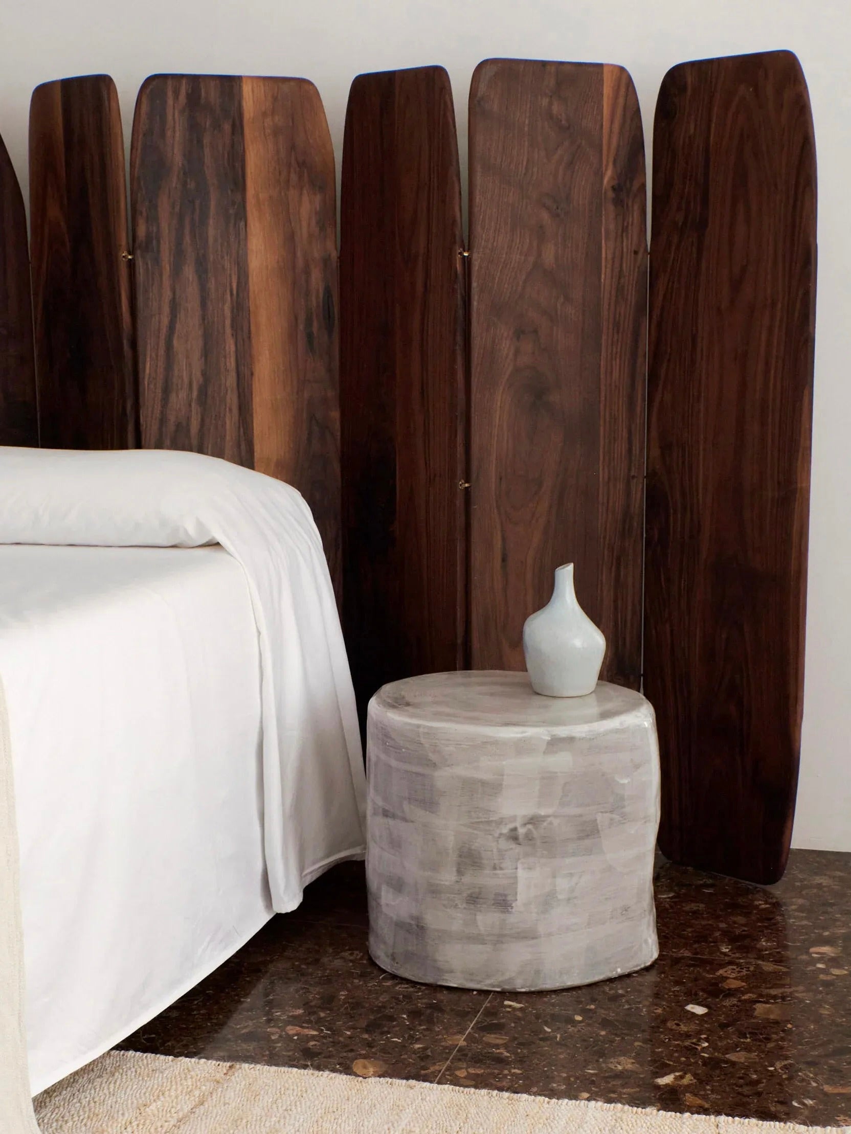 A cozy bedroom corner featuring a bed with white sheets and a wooden headboard consisting of several customizable panels. Beside the bed is a round concrete nightstand with a small, simple white vase on it. The minimalist and serene aesthetic of the room is beautifully enhanced by the addition of Project 213A's "Madeira" Room Divider (small), which complements the solid walnut accents.