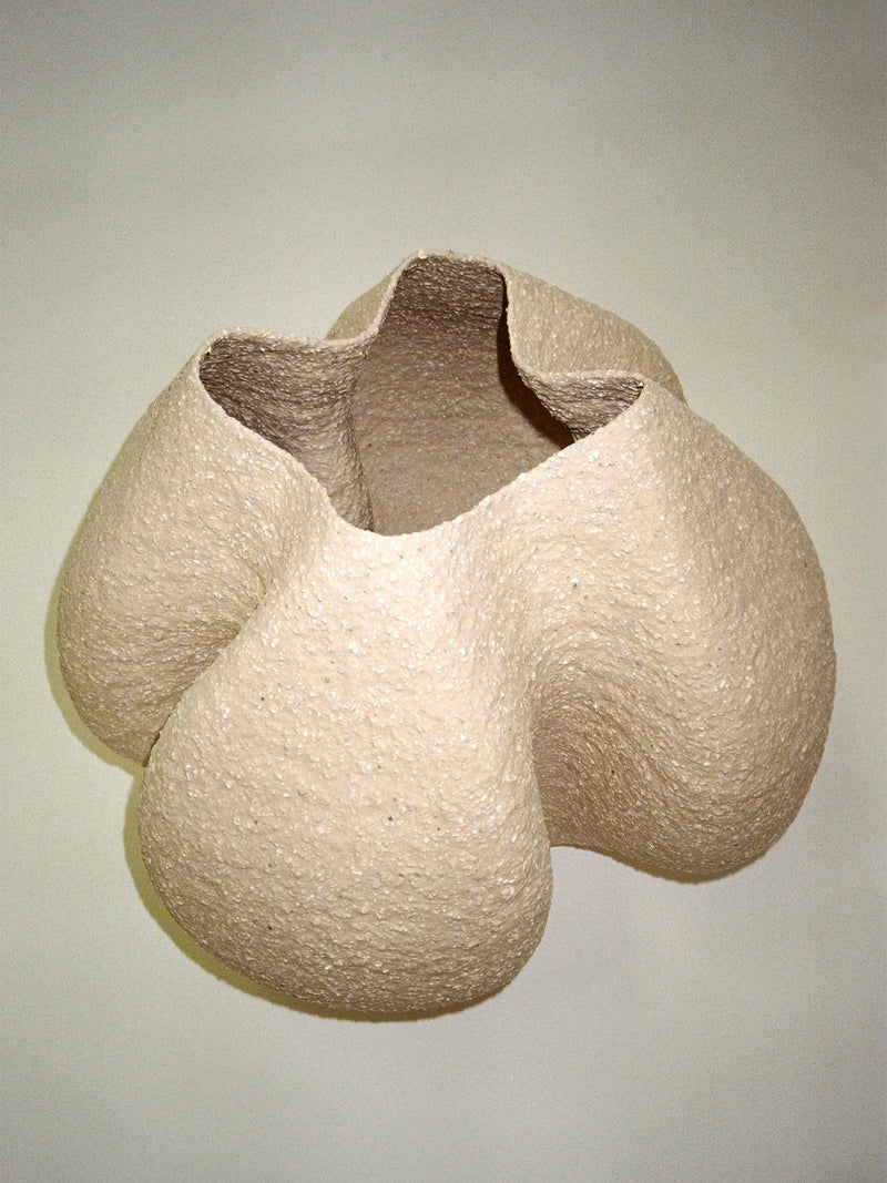 The Growing Vase n.6 by Aiste Bagdonaite is a cream-colored, textured stoneware piece featuring four rounded, bulging sides and an irregular open top, shown against a plain background.