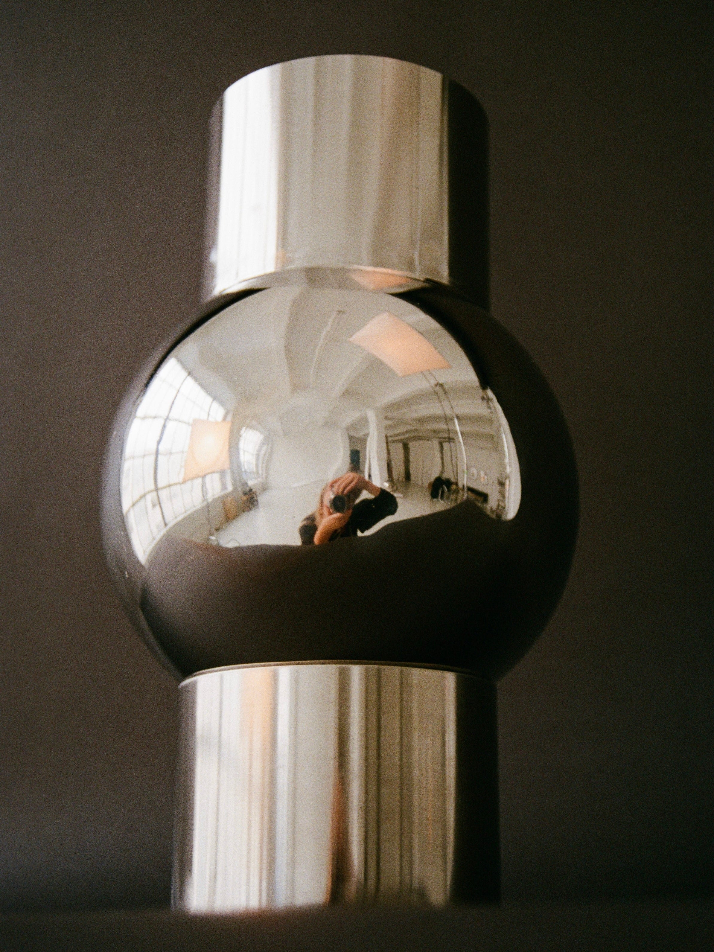 Placed against a dark background is a sleek, cylindrical, metallic object with a reflective stainless steel surface. The reflection reveals an interior space with large windows and someone capturing the photo. The object features a central spherical section flanked by two cylindrical ends, resembling the modern Mica Vase by MUHLY.