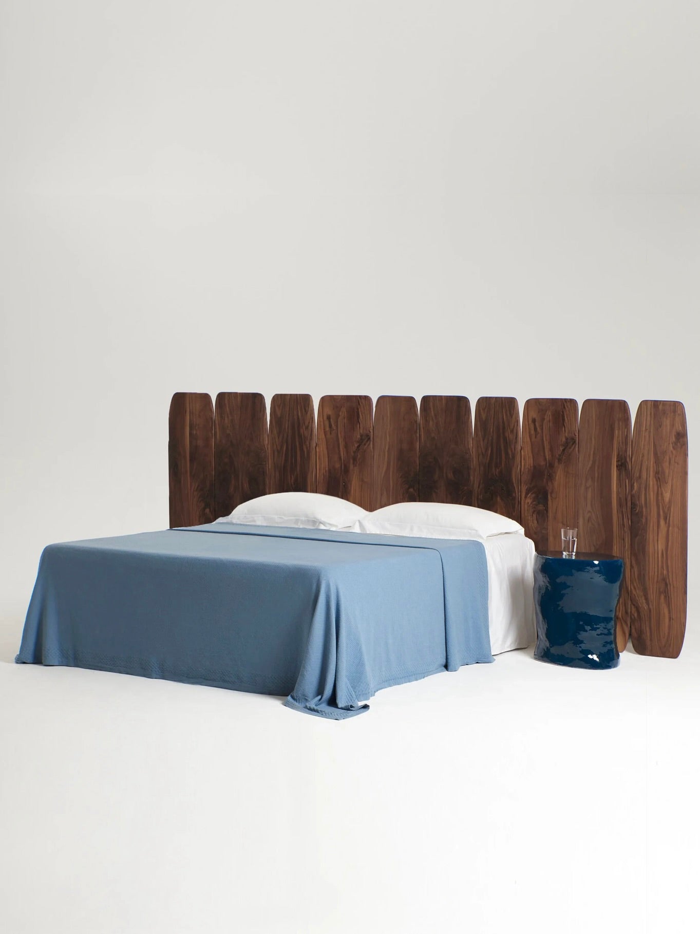 A king-size bed featuring the Madeira Walnut Headboard Large by Project 213A, handmade in Portugal, is adorned with light blue bedding. Two white pillows rest at the head of the bed, while a small blue ceramic side table sits to its right, all set against a plain white background.