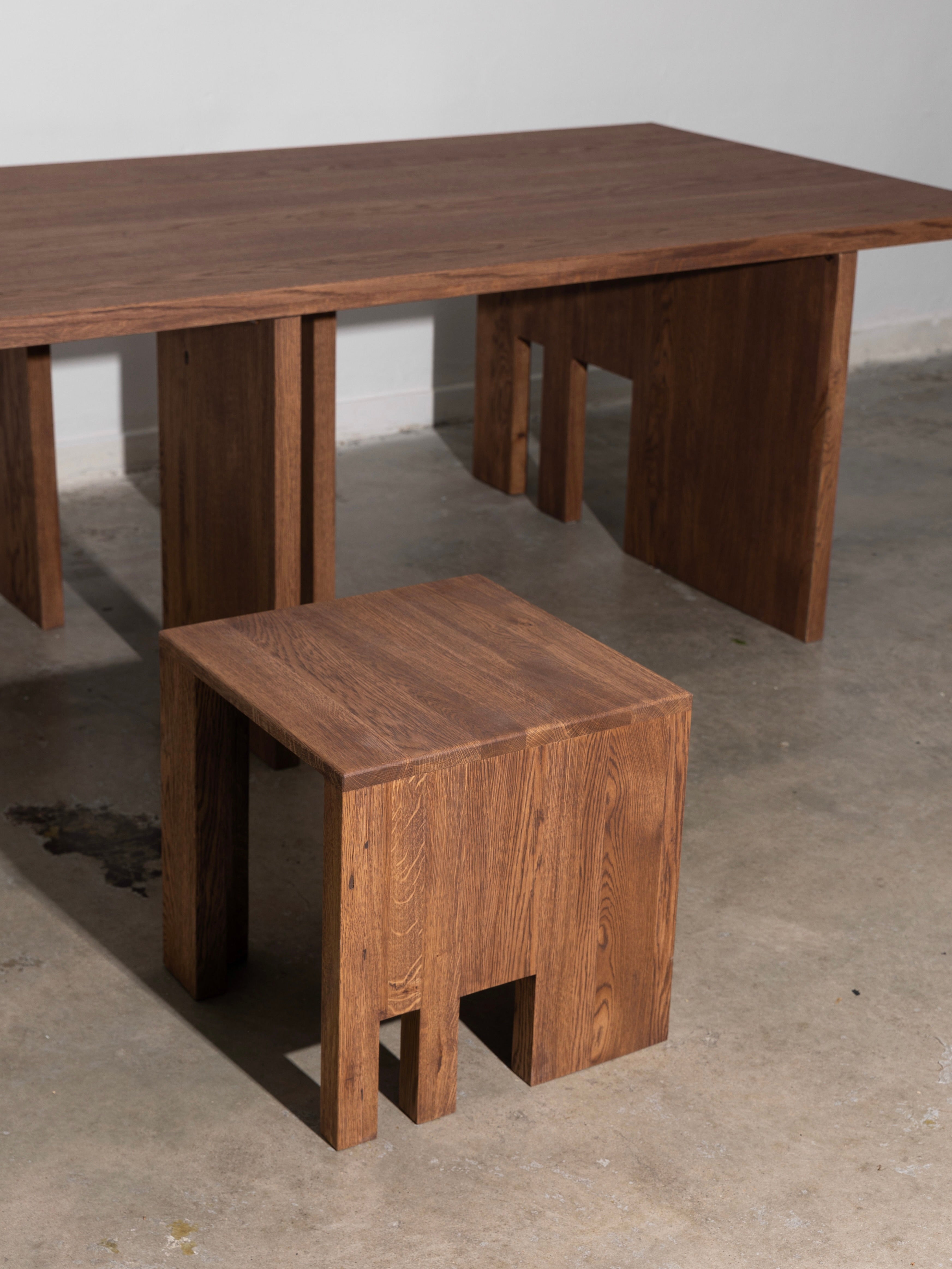 A wooden table and "Dente" Oak Wood Stool from The Good Living&Co., both showcasing exquisite craftsmanship with minimalist design, angular lines, and natural wood grain finish, are set on a light concrete floor.