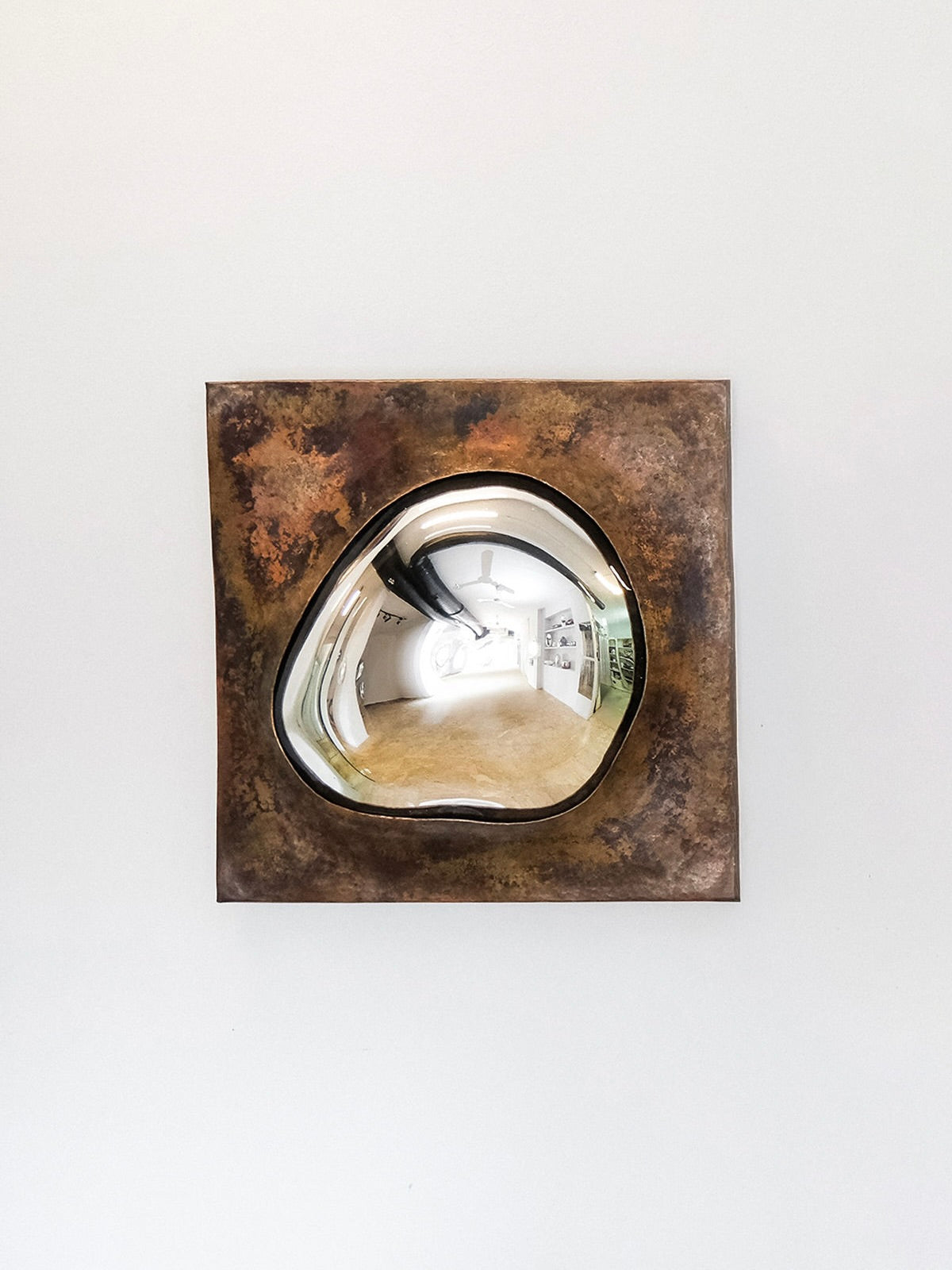 The 'Drop Of Silver On Copper III' by HOMA is a square, rust-colored metal sculpture featuring a copper frame and concave, polished silver center that reflects a distorted view of a white, minimalistic room with partial wooden flooring and visible architectural details.