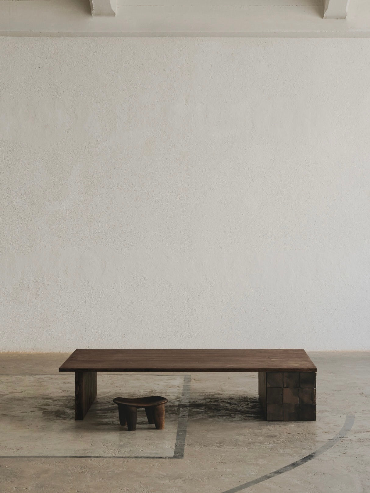 A minimalist room showcases the Cosmos Low Table by ASERIES OF OBSTACLES, SL, and a small stool crafted from European oak against a textured, light-colored wall. The concrete floor's subtle markings complete the unique finish of this serene space.