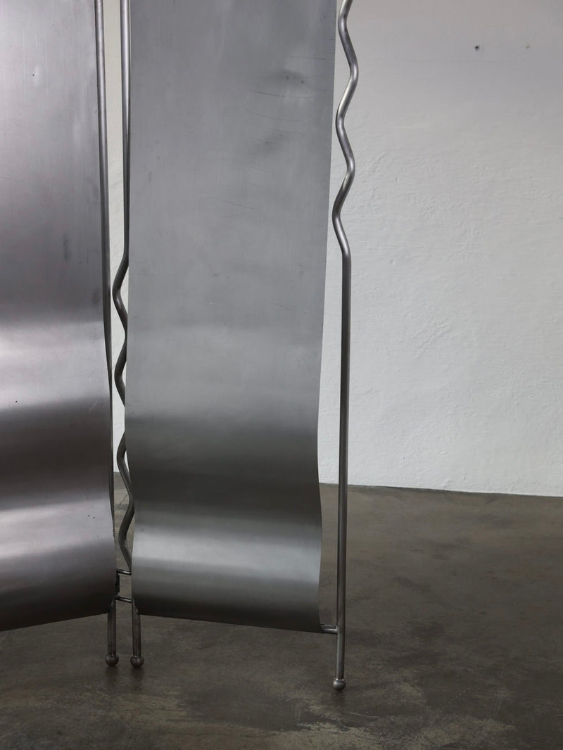 The "Alto" Folding-Screen by The Good Living&Co. is a stainless steel metallic sculpture with a smooth, curved panel and wavy rod, set against a white wall and concrete floor. Crafted in Poland, its form evokes modern art or design.