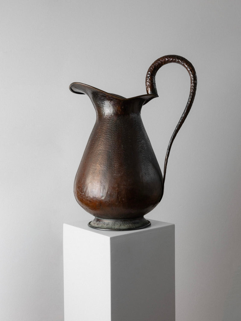 A tall, elegant piece from Bottega Jacobs—a Copper Handmade Jug/Vase from 1950s Italy—stands on a white pedestal against a gray background. Its curved spout and handle with a textured surface evoke vintage charm, adding an antique flair to the scene.