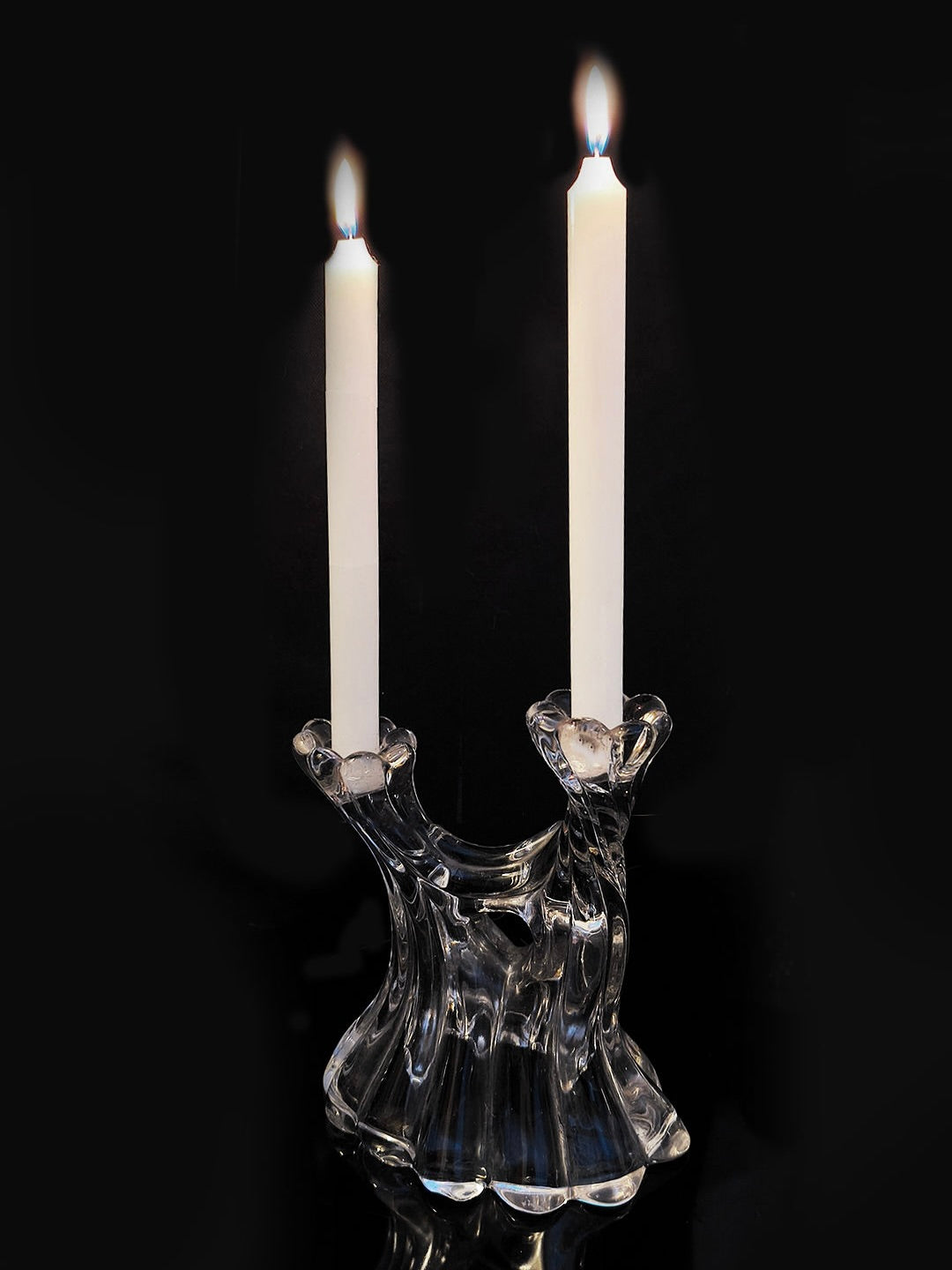 A sophisticated Les Objoies Glass Candle Holder with asymmetrical curves holds two lit white candles, all set against a black background.