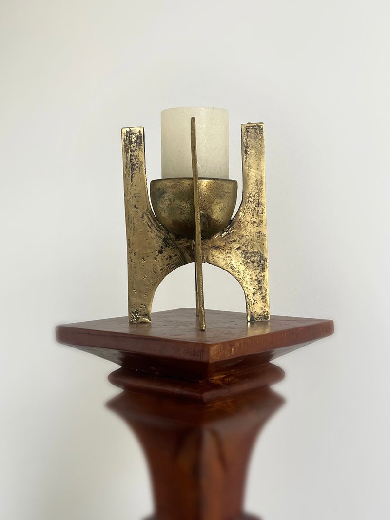A white cylindrical candle sits in the Porter Studio Brutalist Brass Candle Holder, featuring a sculptural abstract design in rustic gold metal on a polished wooden pedestal against a plain white background, adding textured elegance to the scene.
