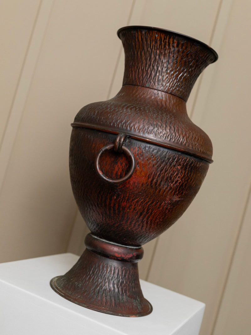 A large, textured decorative piece reminiscent of the Vintage Copper Jug Italy 1960s by Bottega Jacobs, featuring a ring handle on one side, is displayed on a white pedestal against a beige wall. This bronze urn boasts a wide opening at the top and a flared base.