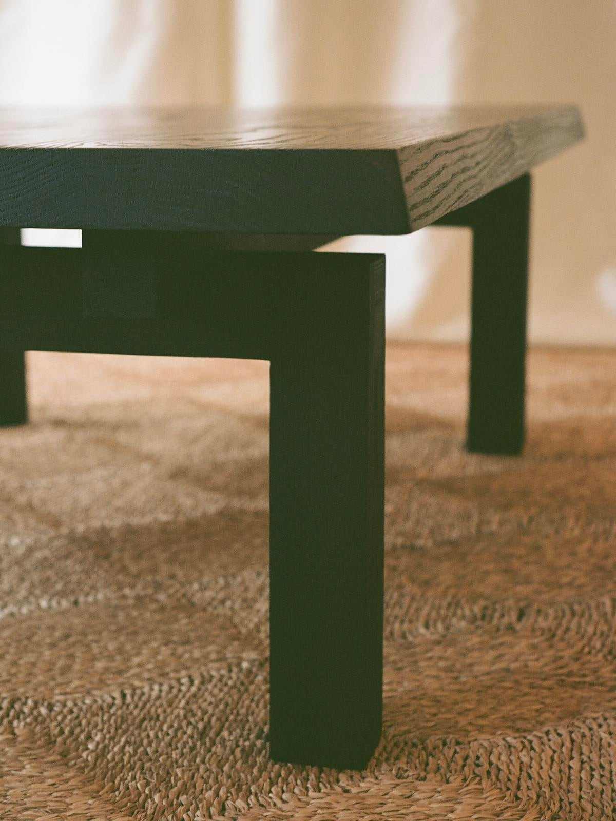 A close-up of the SWM STUDIO Lantern Coffee Table highlights its modern, dark wooden design with a thick tabletop and sturdy legs. This solid oak piece rests on a textured woven rug, embodying Japanese aesthetics with its warm and minimalist atmosphere.