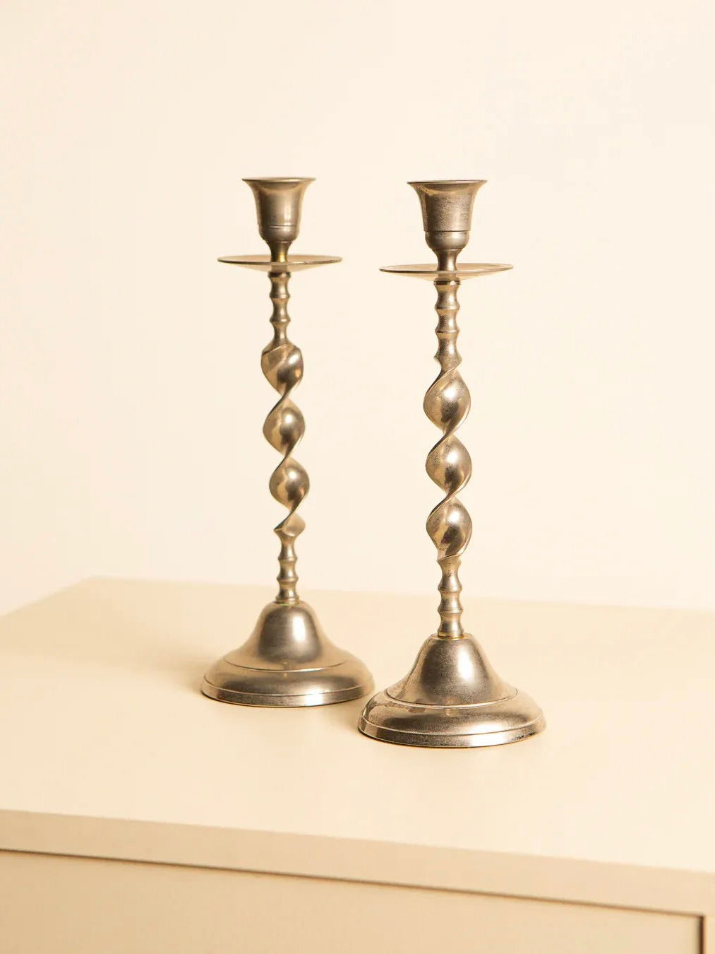 A pair of Treaptyque's 60's Italian Metal Candleholders, featuring twisted, spiral stems, are displayed side by side on a beige surface against a neutral backdrop.