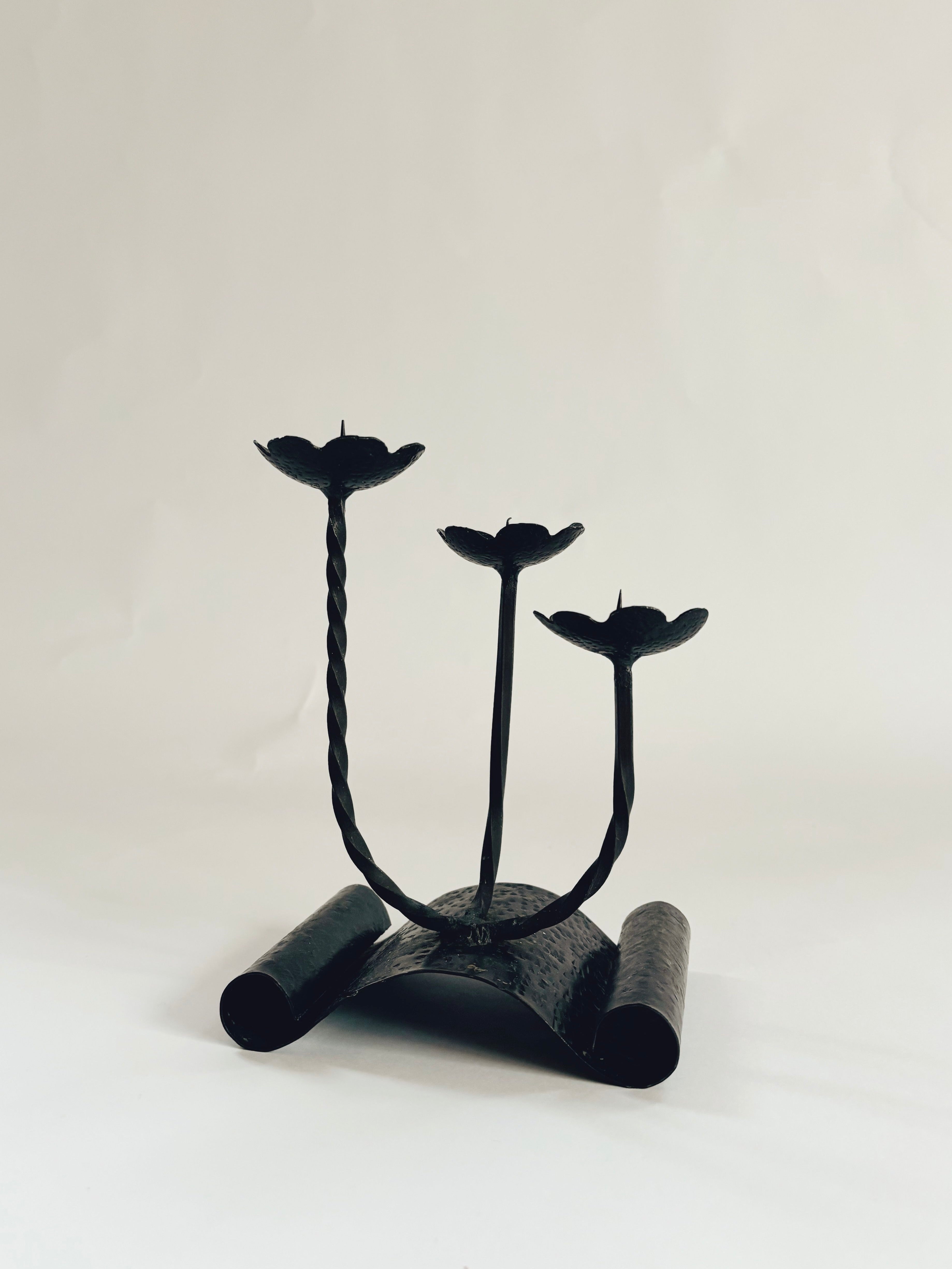 The Handmade Metal Flower Candlestick by Huerto showcases a vintage design with a twisted stem and three flower-shaped platforms for candles, all elegantly standing on a curved, textured base against a plain background.