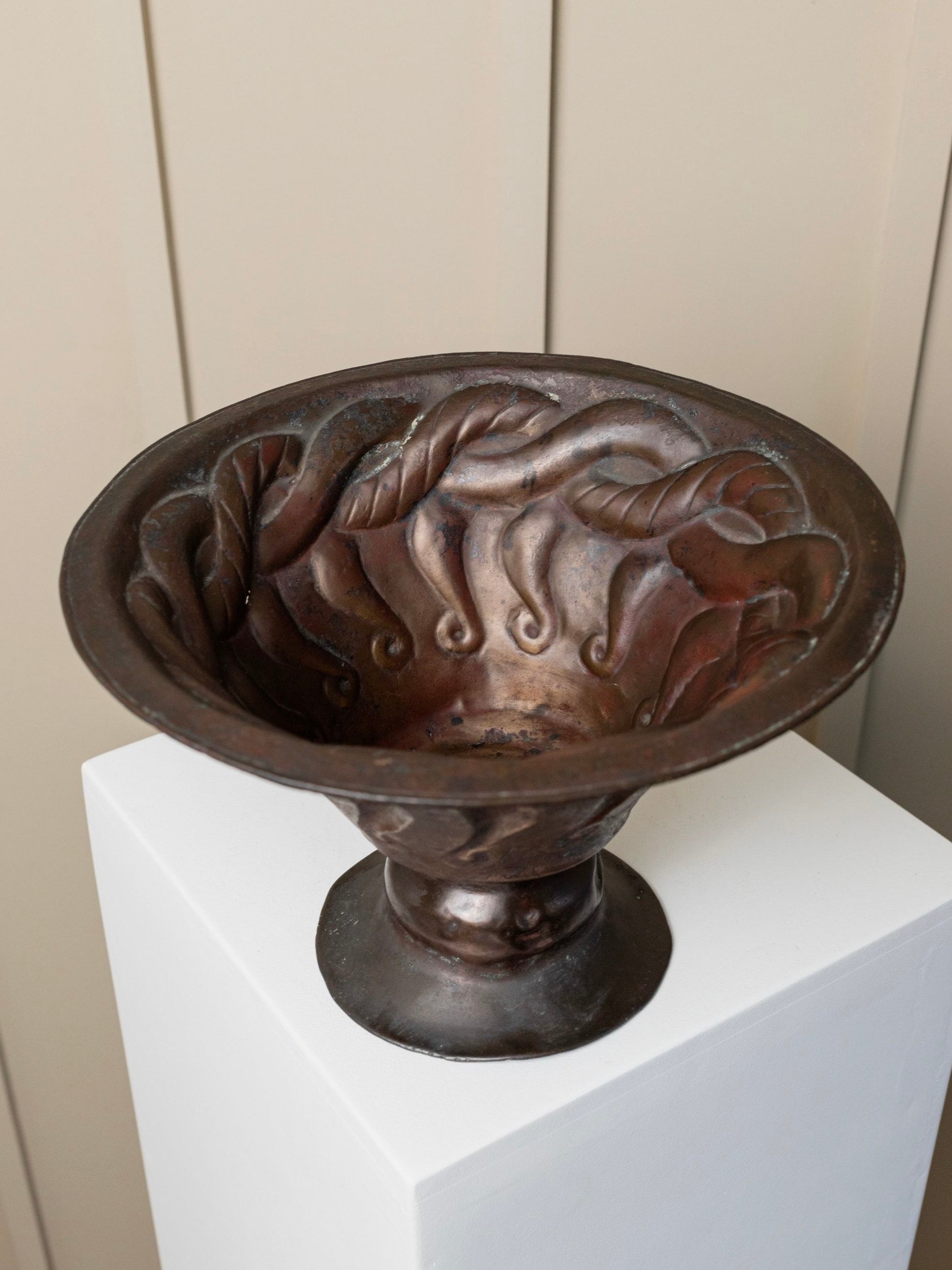 An Antique Fruit Bowl - Hand-Carved Copper by Bottega Jacobs, boasting intricate swirling designs and a vintage patina, sits elegantly on a white pedestal against a neutral-colored wall.