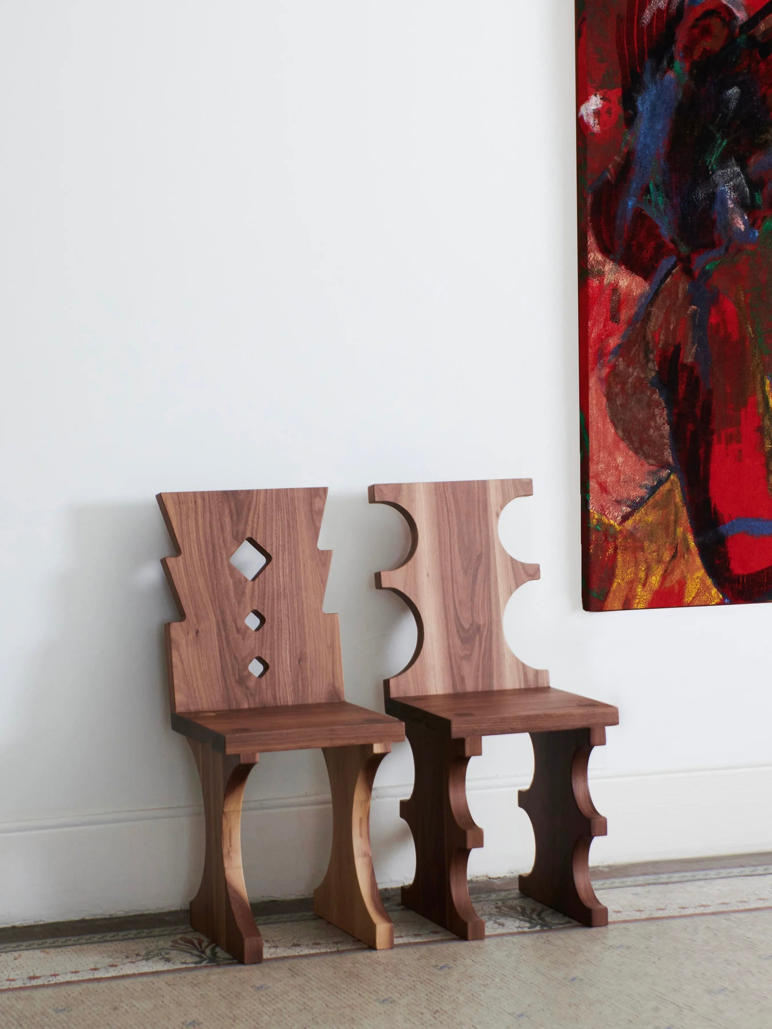 Two uniquely designed Portugal Chair No 7 pieces crafted from solid walnut by Project 213A, featuring abstract, geometric shapes meticulously carved by skilled artisans, are placed against a white wall. To the right, part of a colorful, abstract painting is visible, partially cropped out by the edge of the image.