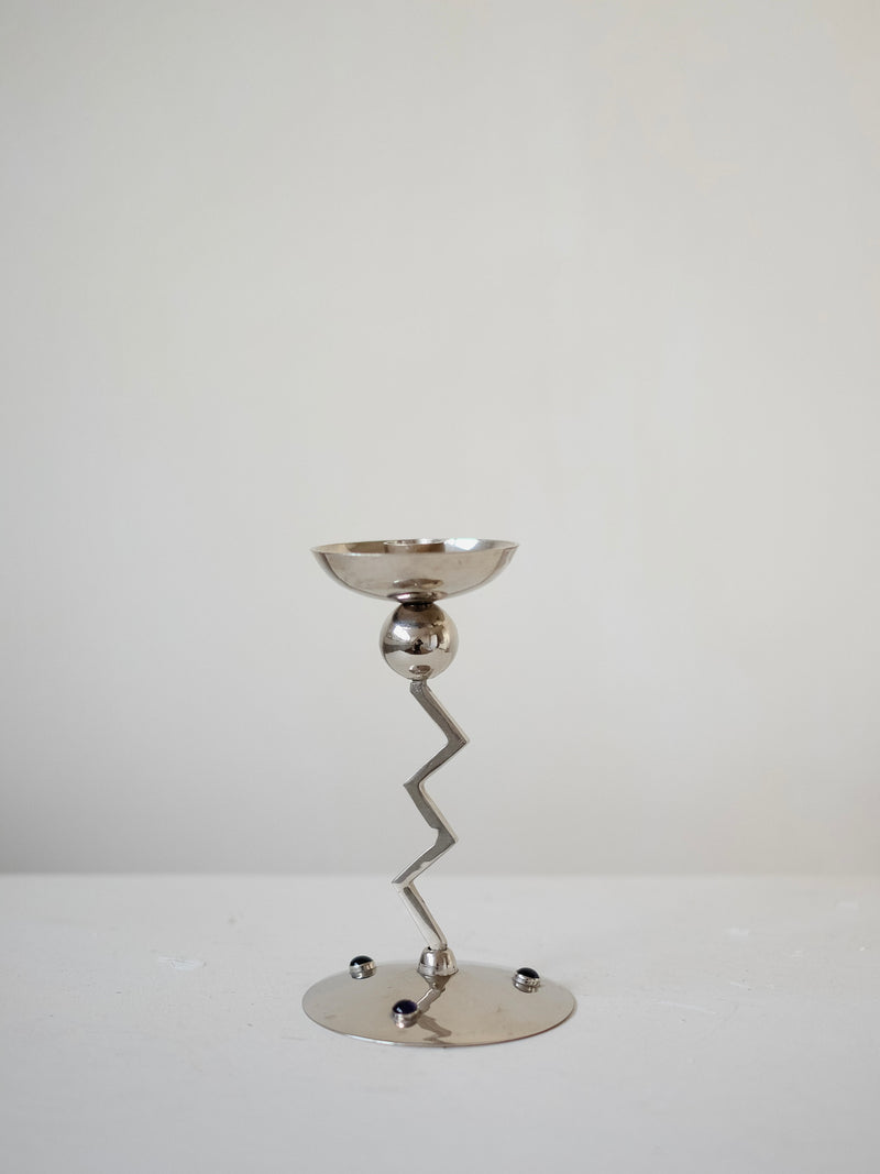 The Zigzag Candleholder by septembre studios, featuring a modern silver design with a zigzag stem, circular base, and decorative spheres, stands against a plain white background.
