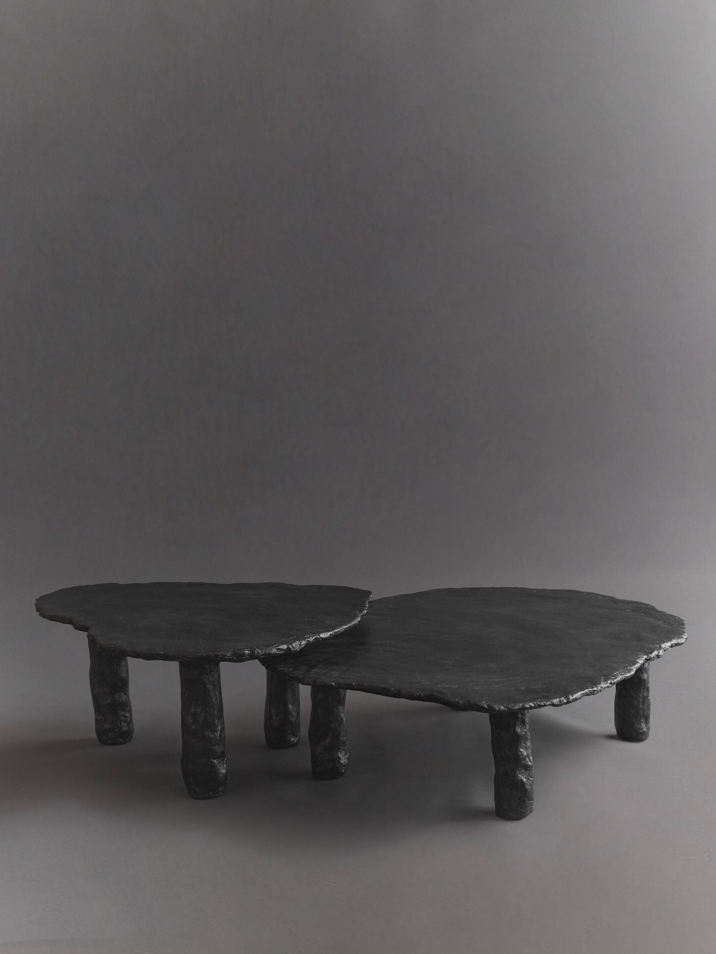 The Roca Nested Coffee Tables by Ombia Studio are two matte black, handmade pieces with abstract, irregular shapes. They feature uneven legs against a gray background, showcasing overlapping designs in varying heights and a rough, textured finish.