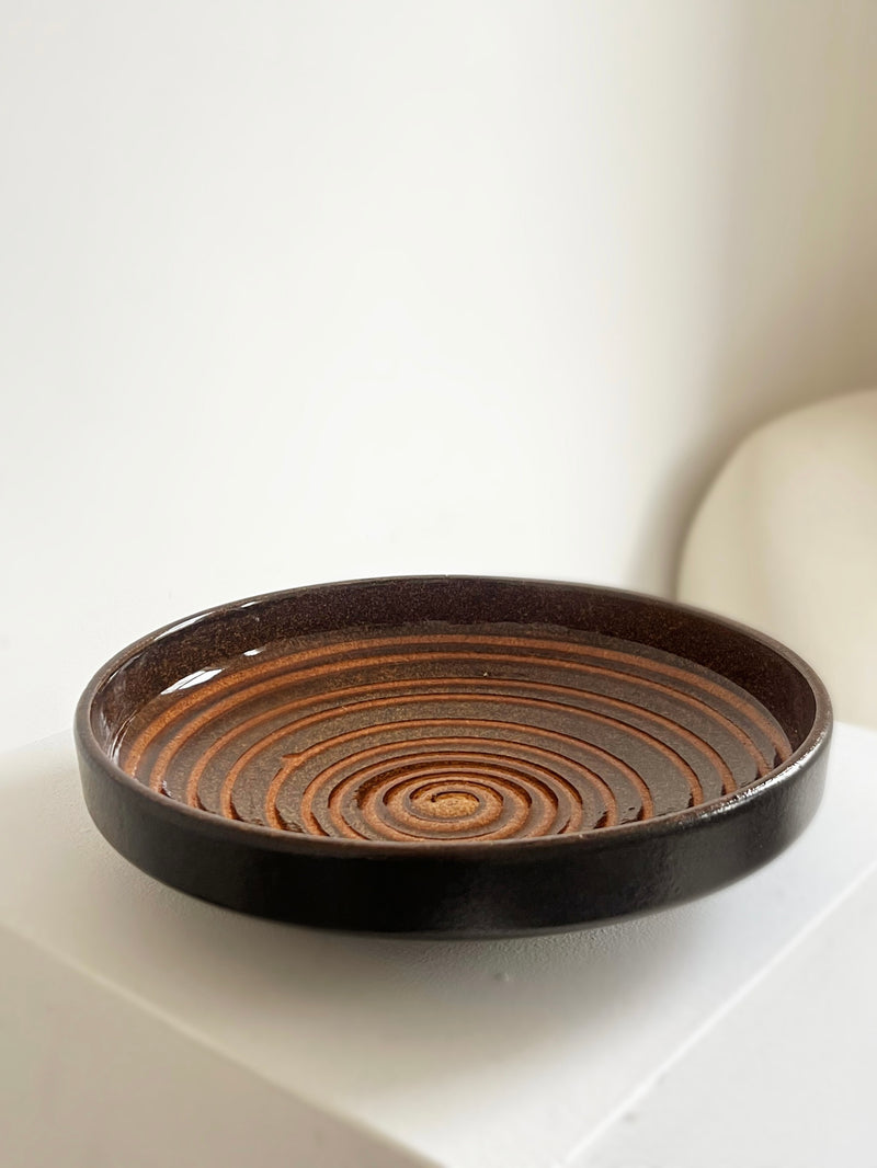 The Spiral Ceramic Dish by Porter Studio, featuring dark brown concentric circular ridges, is elegantly showcased on a white pedestal against a neutral background, making it perfect for sophisticated kitchen use.