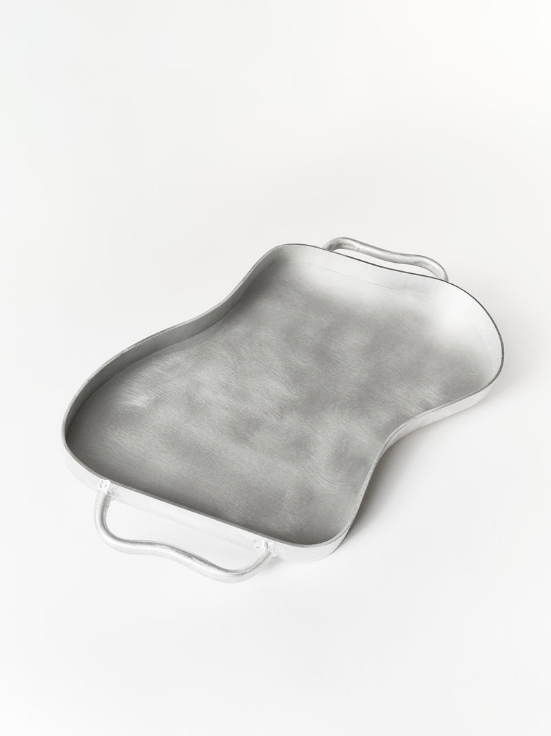 A metallic Serving Tray by Six Dots Design, with curved edges, two handles, an abstract irregular shape, and a smooth brushed texture, handmade in a North London workshop, rests on a plain white surface.