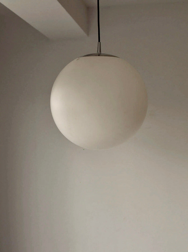 A modern, minimalistic Glass Globe Lamp Peill & Putzler from CU-RA-TED hangs from a black cord against a plain white wall and ceiling. The light fixture has a smooth and sleek design, adding a contemporary touch to the room while retaining its vintage condition charm.